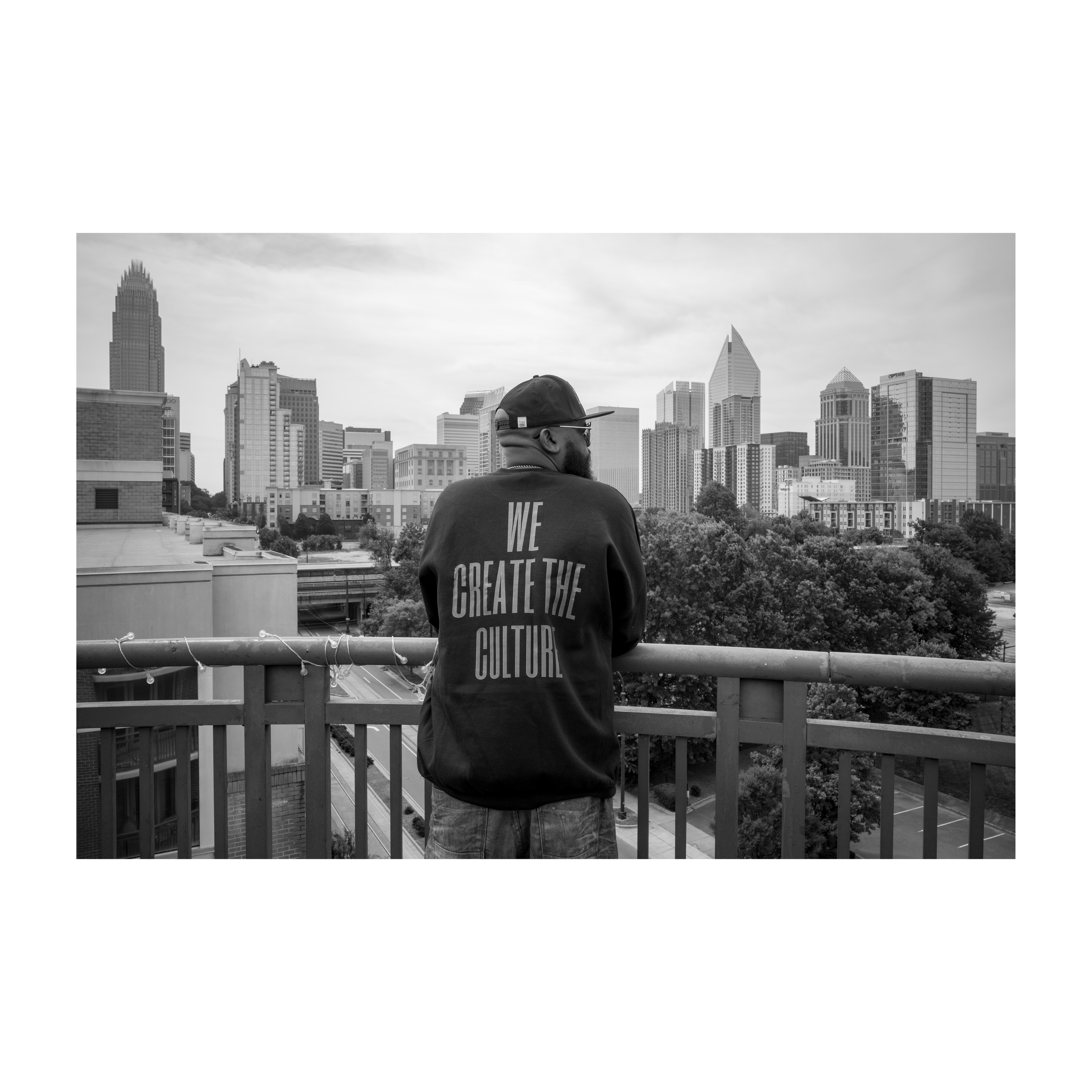 A man wearing a cap and jacket viewed from behind, standing on a balcony overlooking a city skyline with tall buildings.