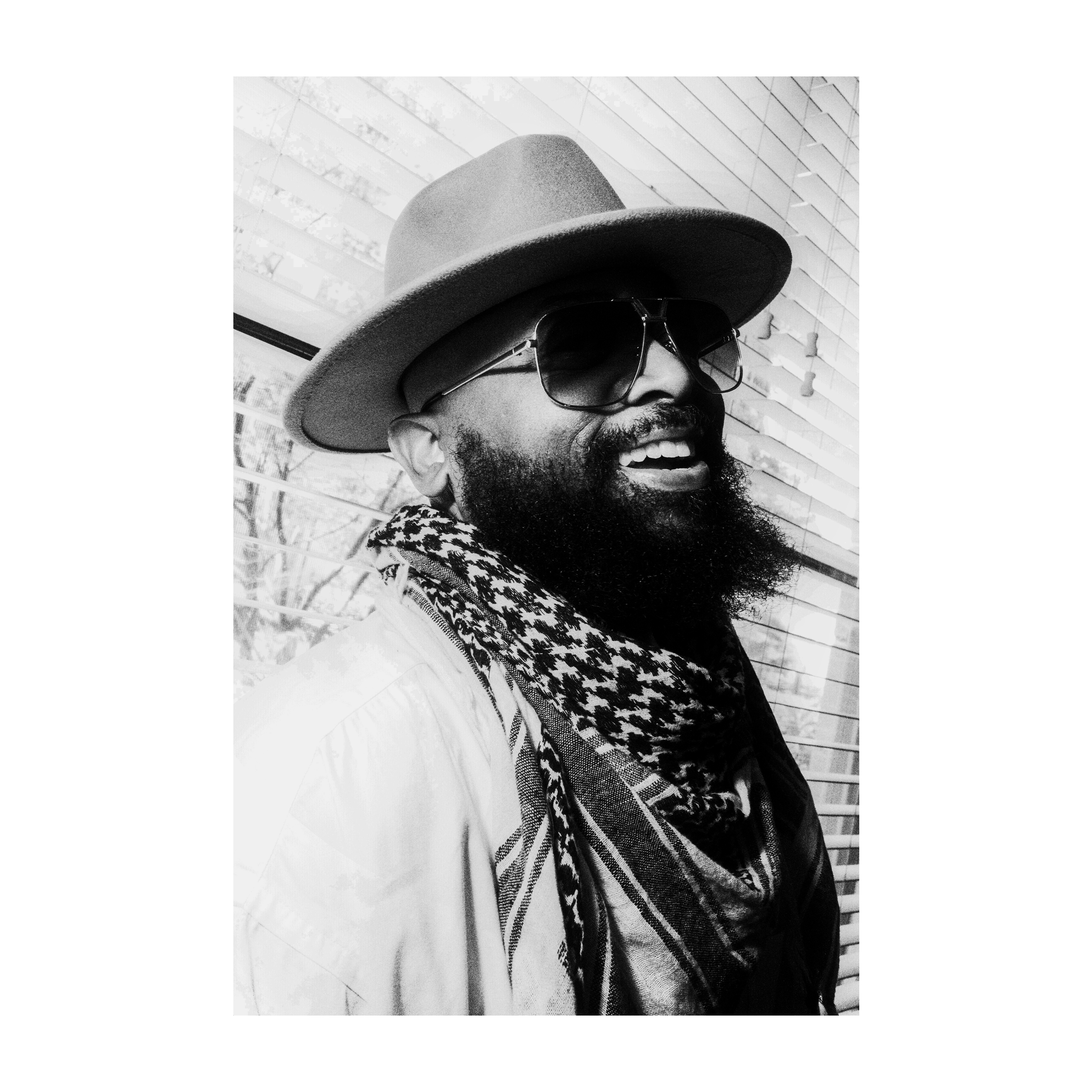 A person smiling, wearing a large hat, sunglasses, and a patterned scarf, sitting outside near a modern building with glass windows.