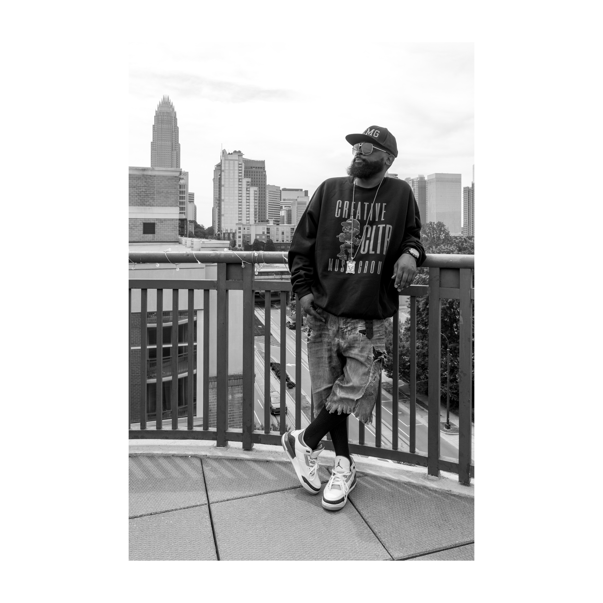 A man with a beard, wearing sunglasses, a baseball cap, and casual clothes, leaning on a railing on a rooftop with a city skyline in the background.