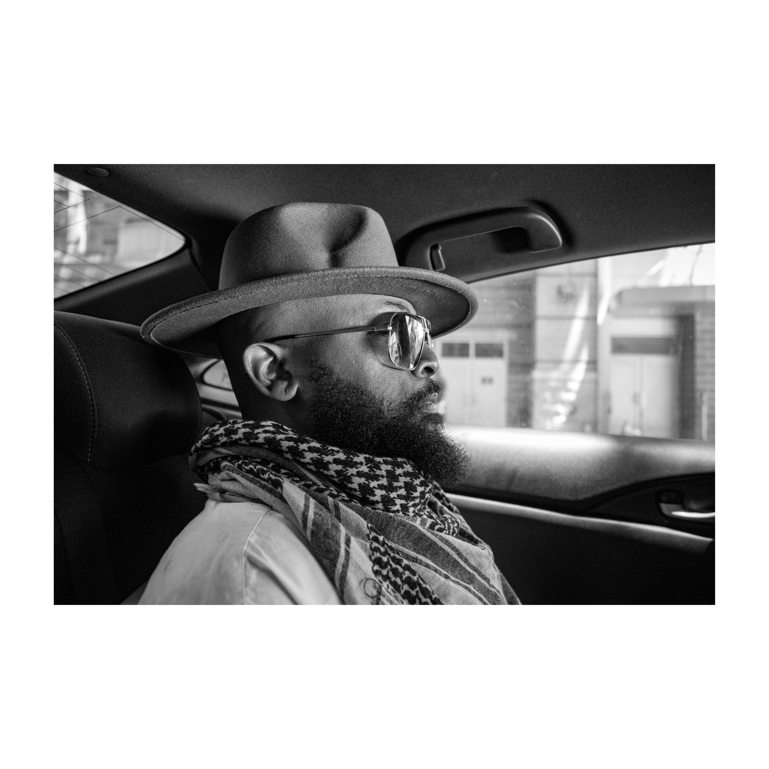 A person with a beard wearing glasses and a hat sits in a vehicle, looking to the right side of the image. The background shows a blurred building and parking structure.