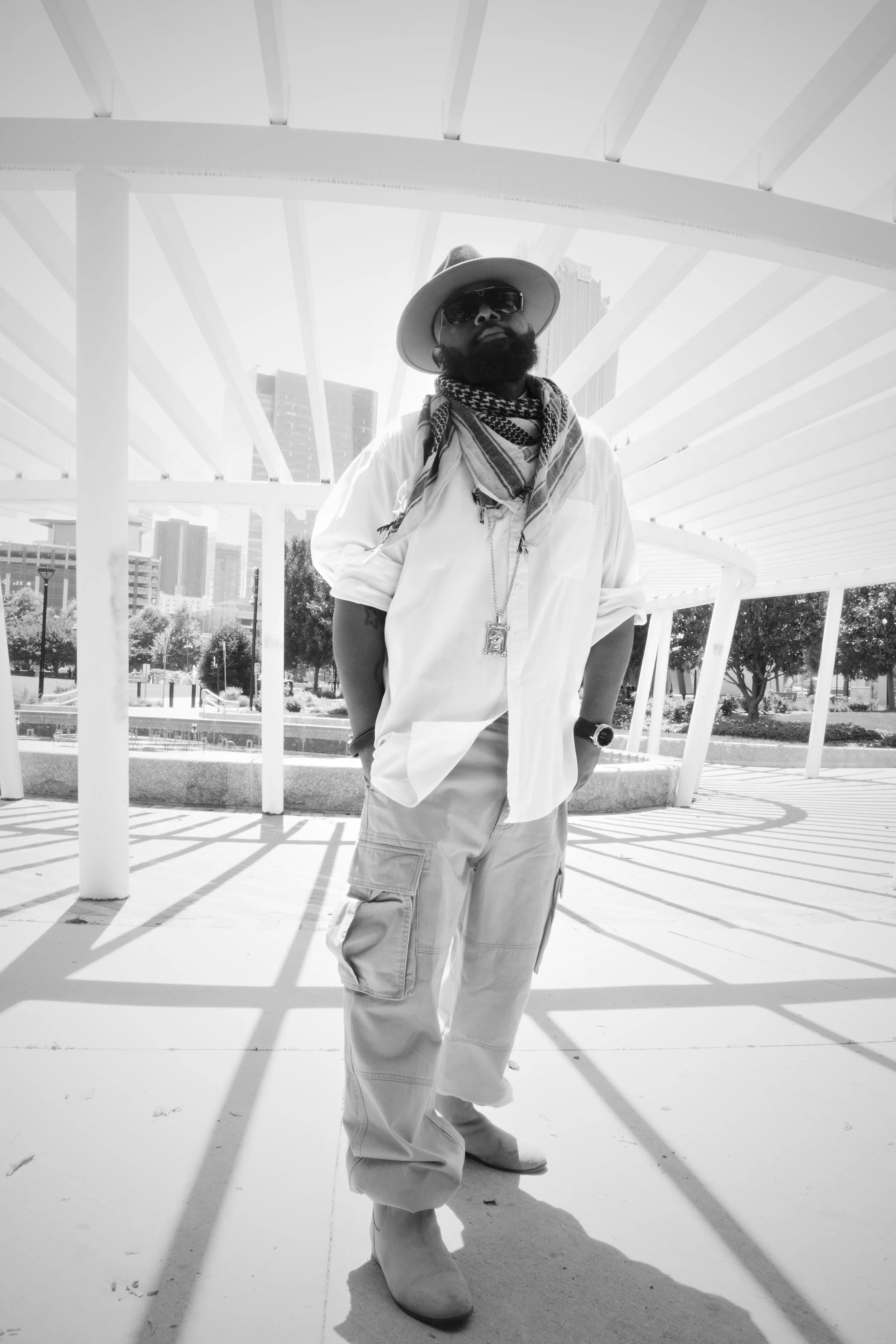 A man wearing a wide-brimmed hat, sunglasses, a scarf around his neck, a long-sleeve shirt, cargo pants, and cowboy boots, standing under a modern architectural structure with a city skyline in the background.