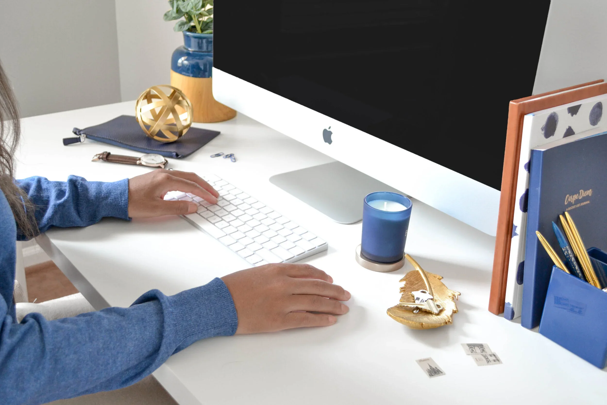 Person working at a white desk with a large desktop computer, a watch, a blue pen holder, a plant, folders, and decorative items.