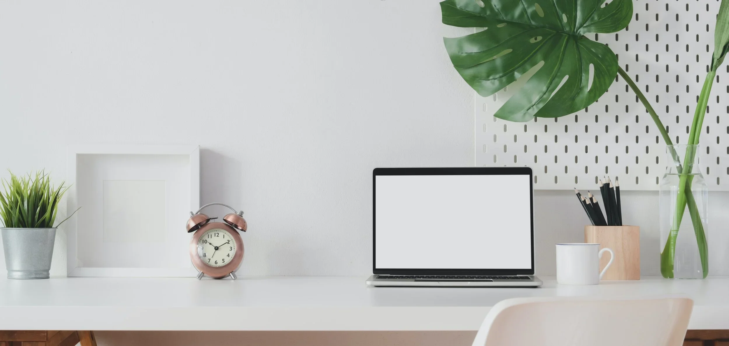 Organized white desk with potted plant, pink alarm clock, open laptop, white mug, pencil holder, and tall vase with monstera leaves against a white wall with a white pegboard.
