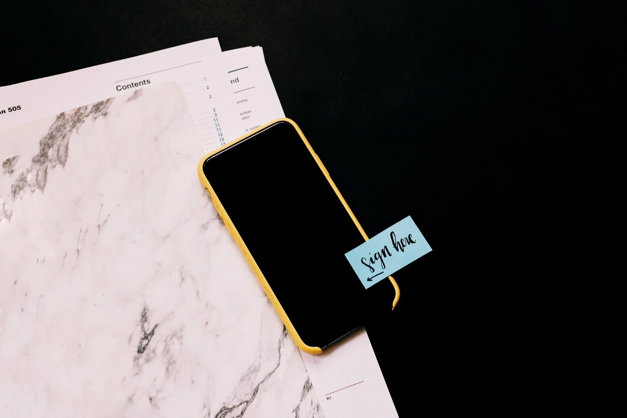 A smartphone with a yellow case displaying a sign that says 'sign here,' resting on top of a blue sticky note, alongside a marble-textured paper with a table of contents and a map, on a black surface.