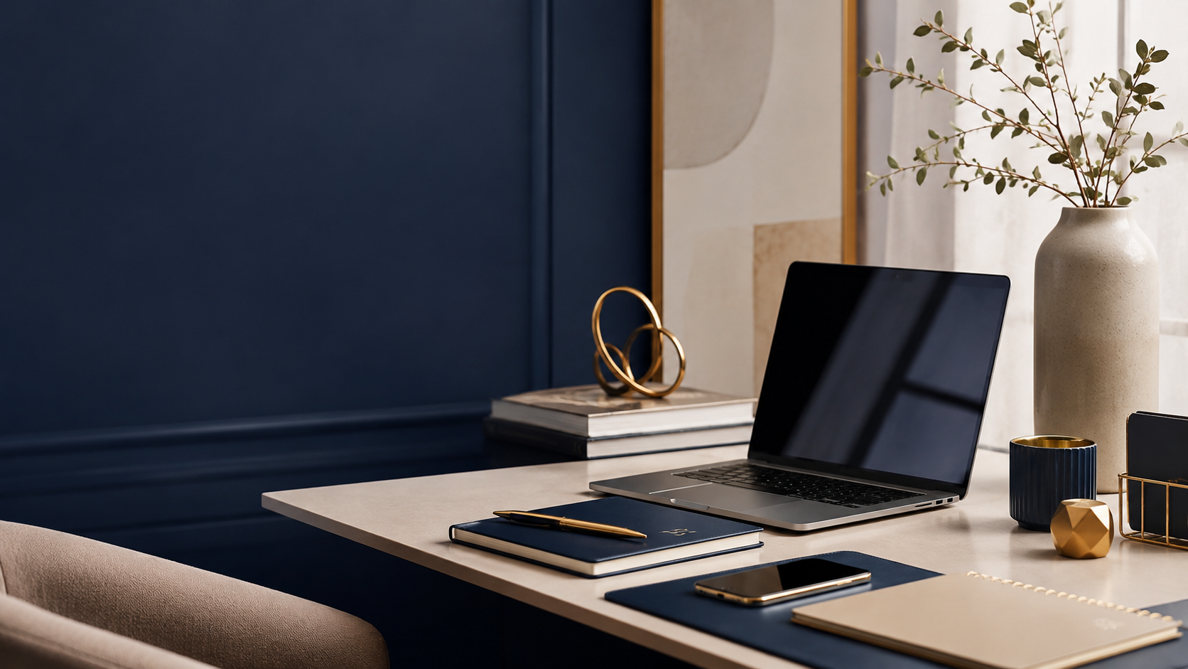 A modern workspace with a laptop, notebooks, and accessories on a desk near a vase with greenery, with a navy blue wall and abstract artwork in the background.