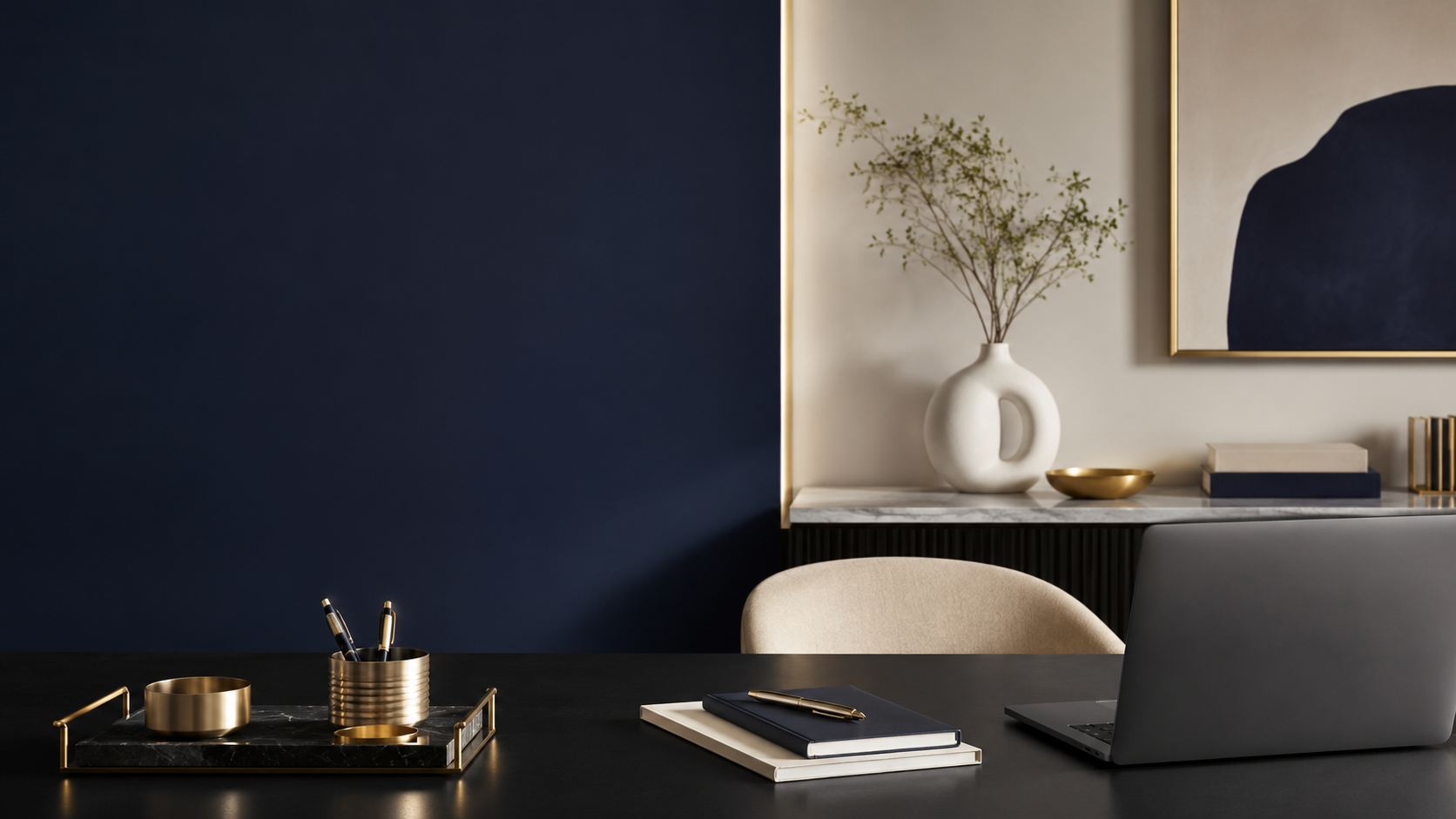 Modern workspace with a black desk, notebooks, pen, gold desk accessories, a laptop, beige chair, and a sideboard with a white vase with green branches, gold bowl, and framed abstract art on a dual-tone wall.
