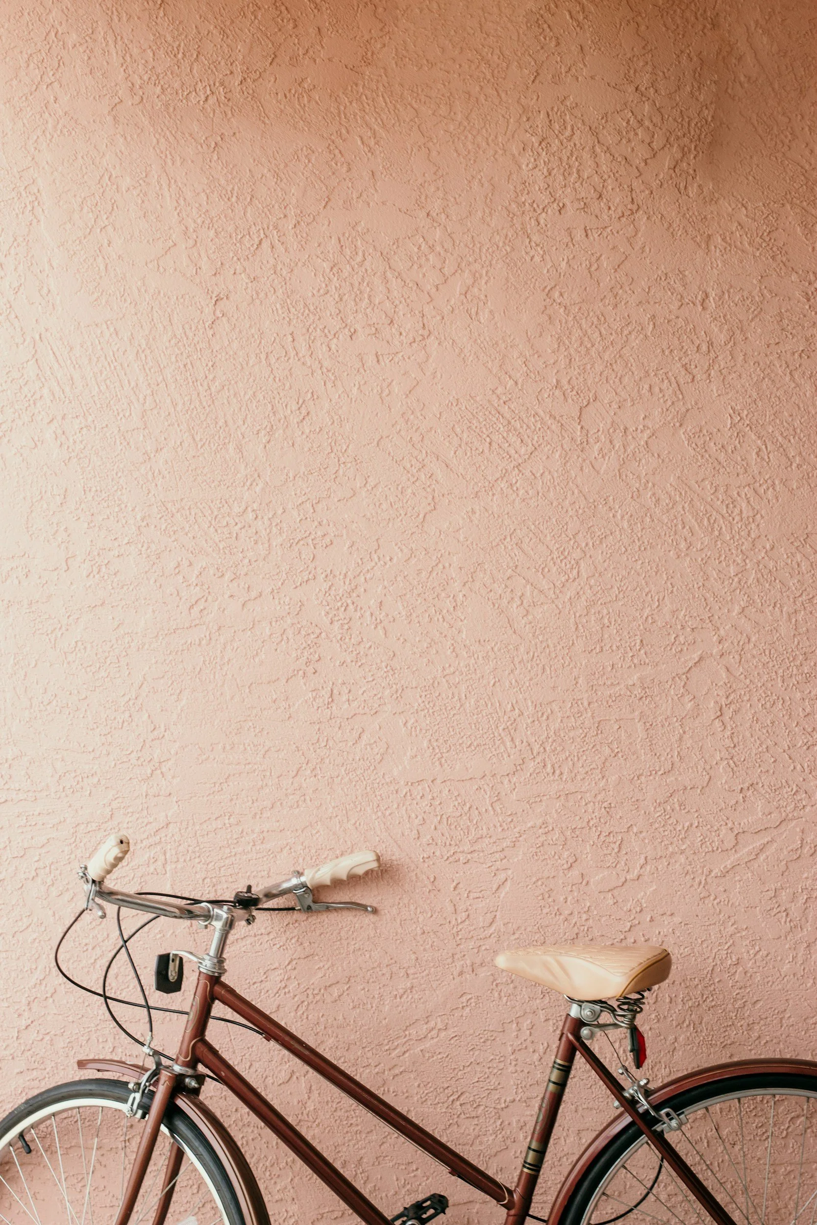 A pink bicycle with beige seat and white handlebars leaning against a pink textured wall.