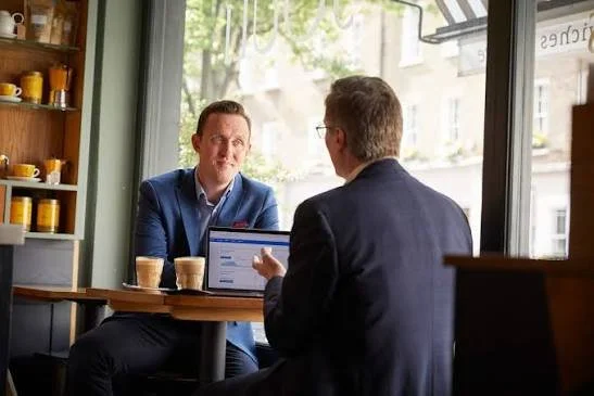 Two men having a conversation in a coffee shop, one using a laptop. The man facing the camera is dressed in a blue suit, while the other has glasses and is wearing a dark blazer.