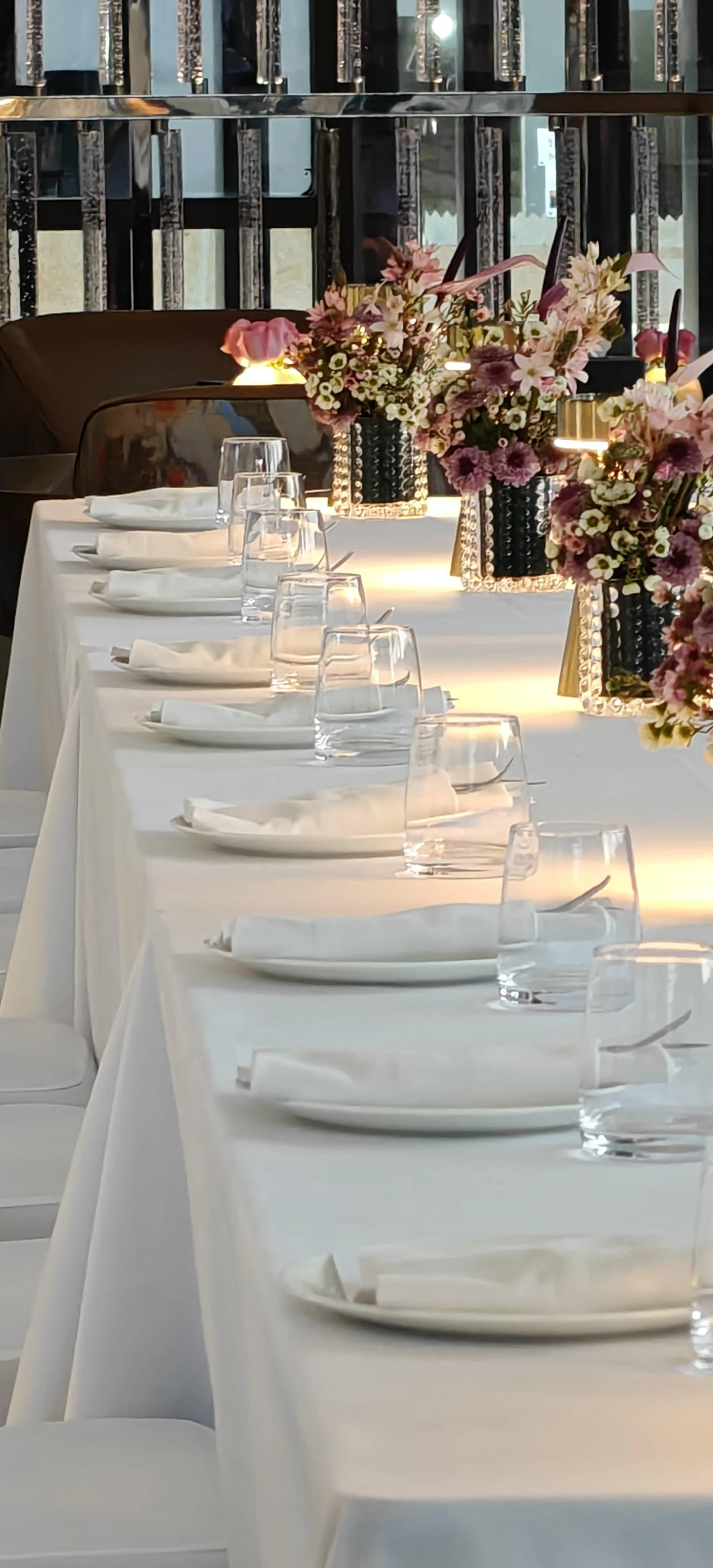 Elegant dining table set with white tablecloth, white plates, clear water glasses, and neatly folded napkins; decorated with pink and purple floral centerpieces in black vases, ambient lighting in a modern room.