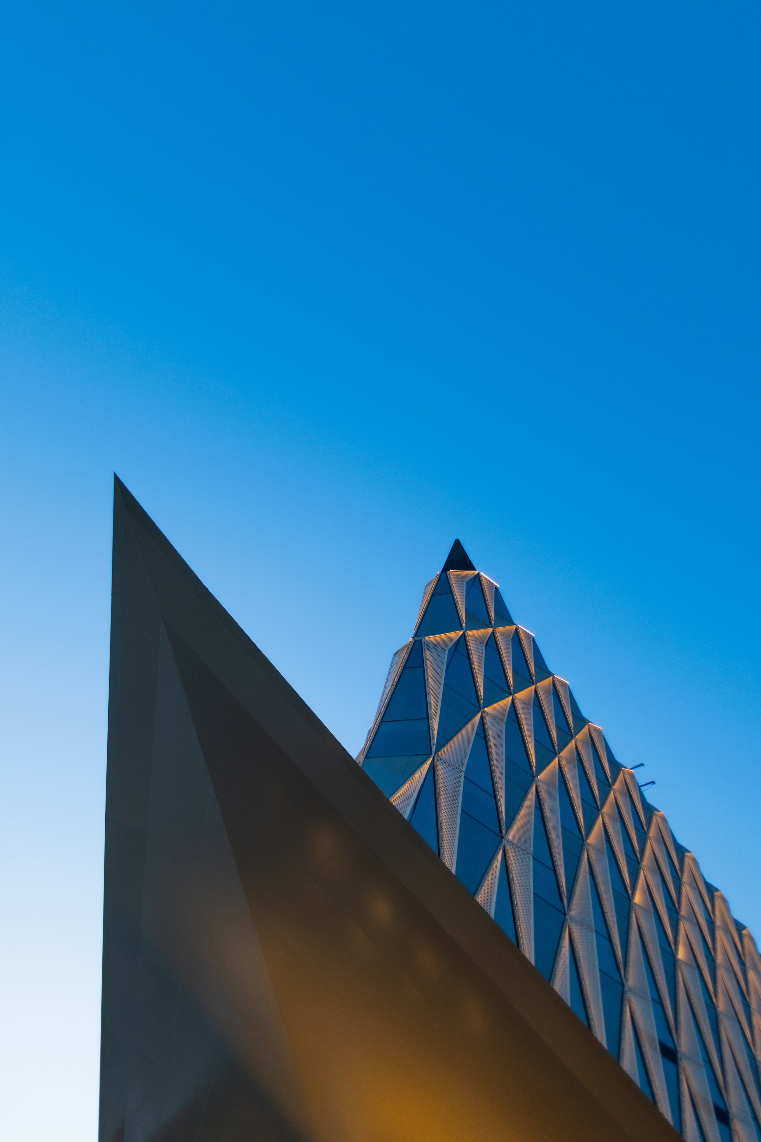 Close-up view of a modern architectural building with a geometric, triangular-patterned facade reflecting city lights against a clear sky.