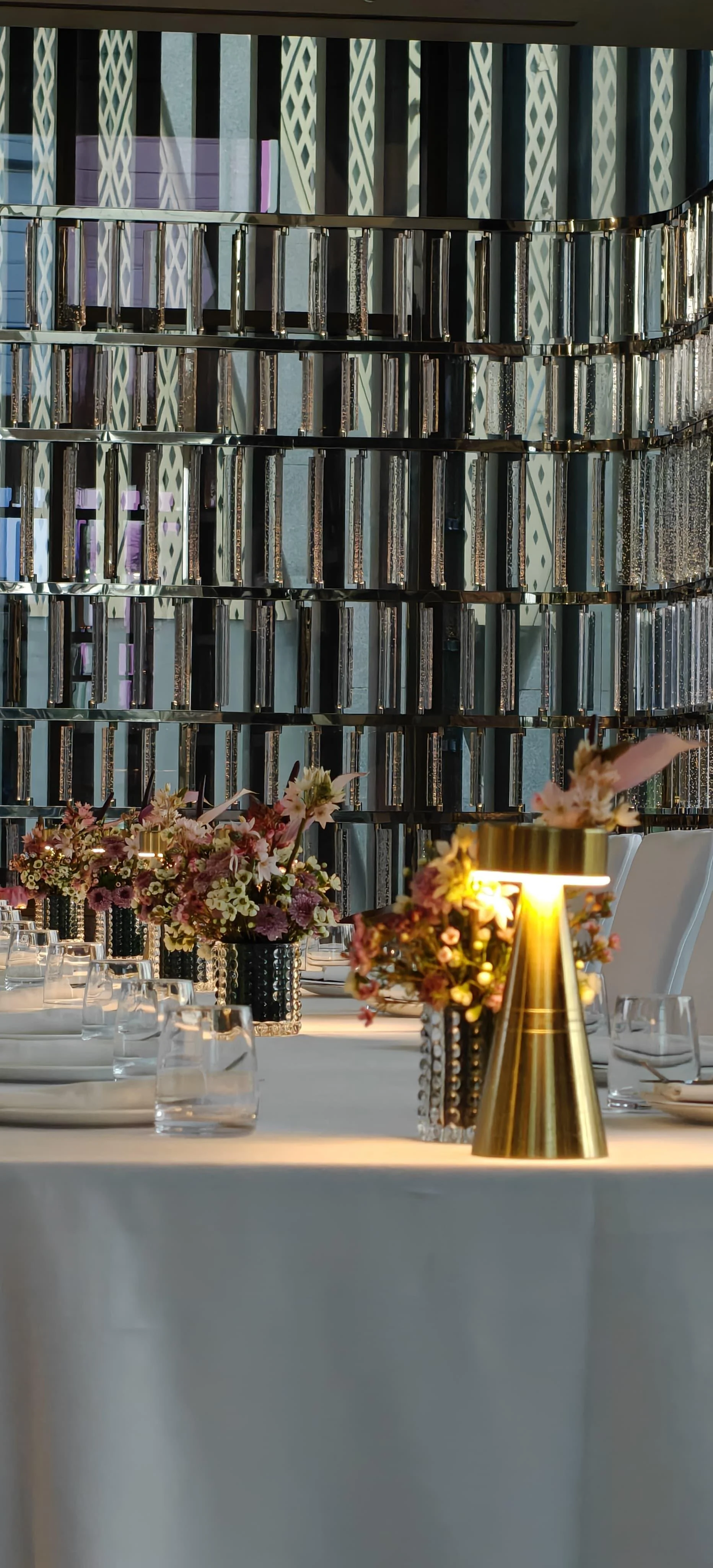 Elegant dining table set with floral centerpieces and modern tableware, with a reflective decorative wall in the background.