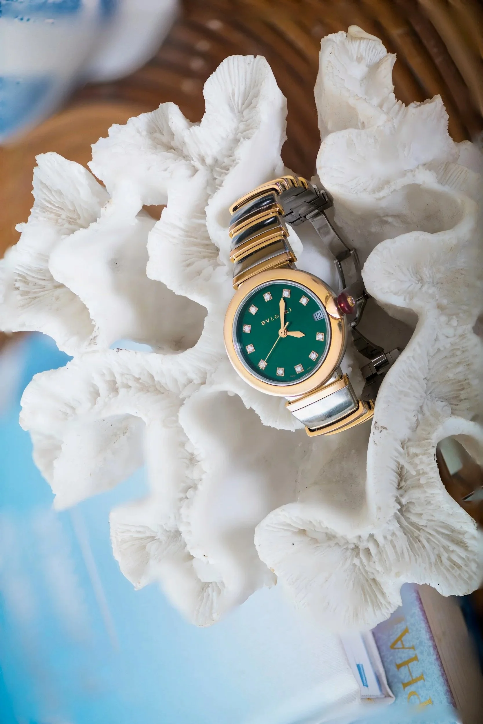 A wristwatch with a green dial and diamond hour markers resting on a white, textured, branched object, possibly coral, with a blurred blue and brown background.