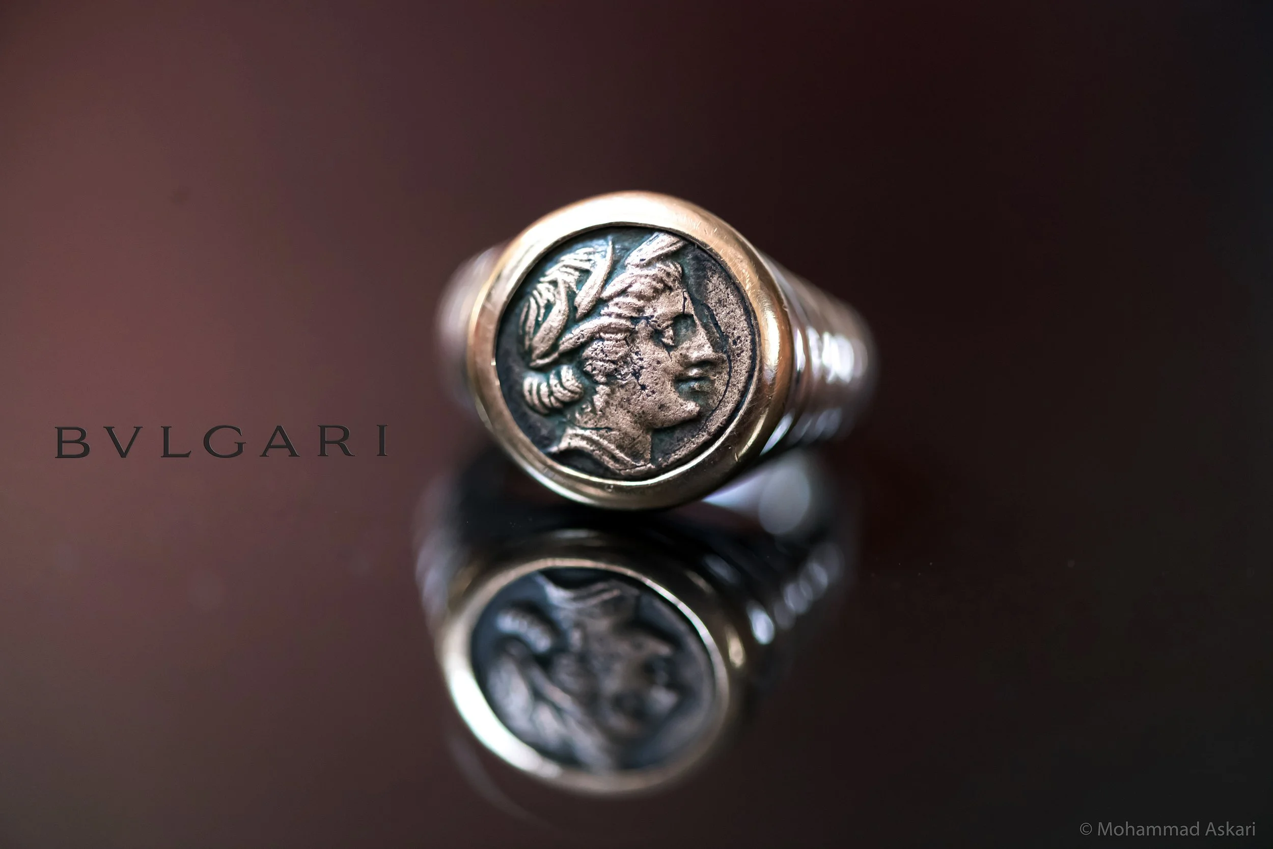 Close-up of a ring with a coin inside it, featuring a profile of a woman with an intricate hairstyle. The background is dark, and the ring appears to be made of metal. The word 'BVLGARI' is visible along the bottom left.