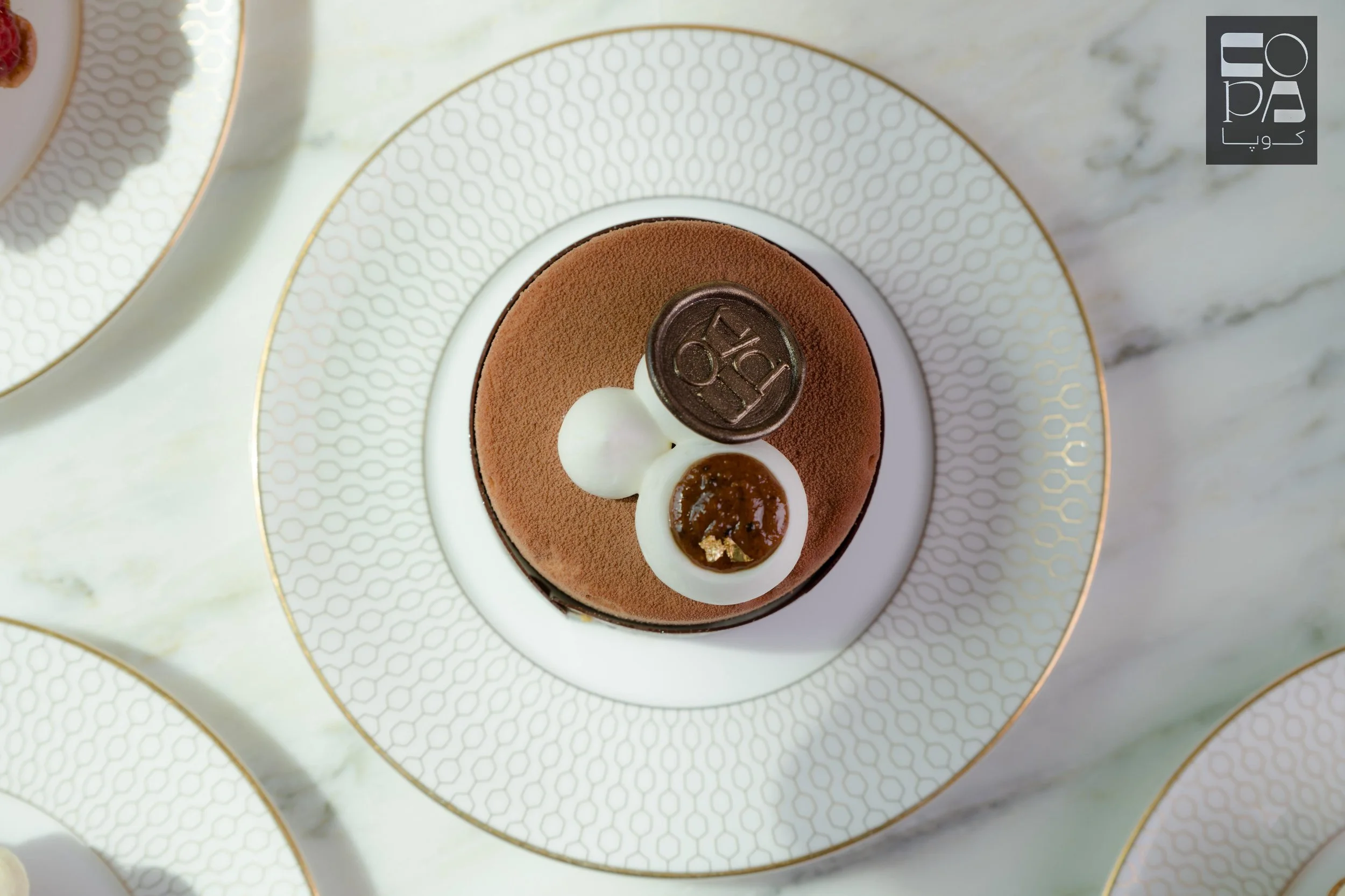 A close-up of an elegant dessert served on a decorative white plate with a gold pattern. The dessert is a small round chocolate mousse topped with a chocolate coin, a dollop of whipped cream, and a caramelized candy piece.