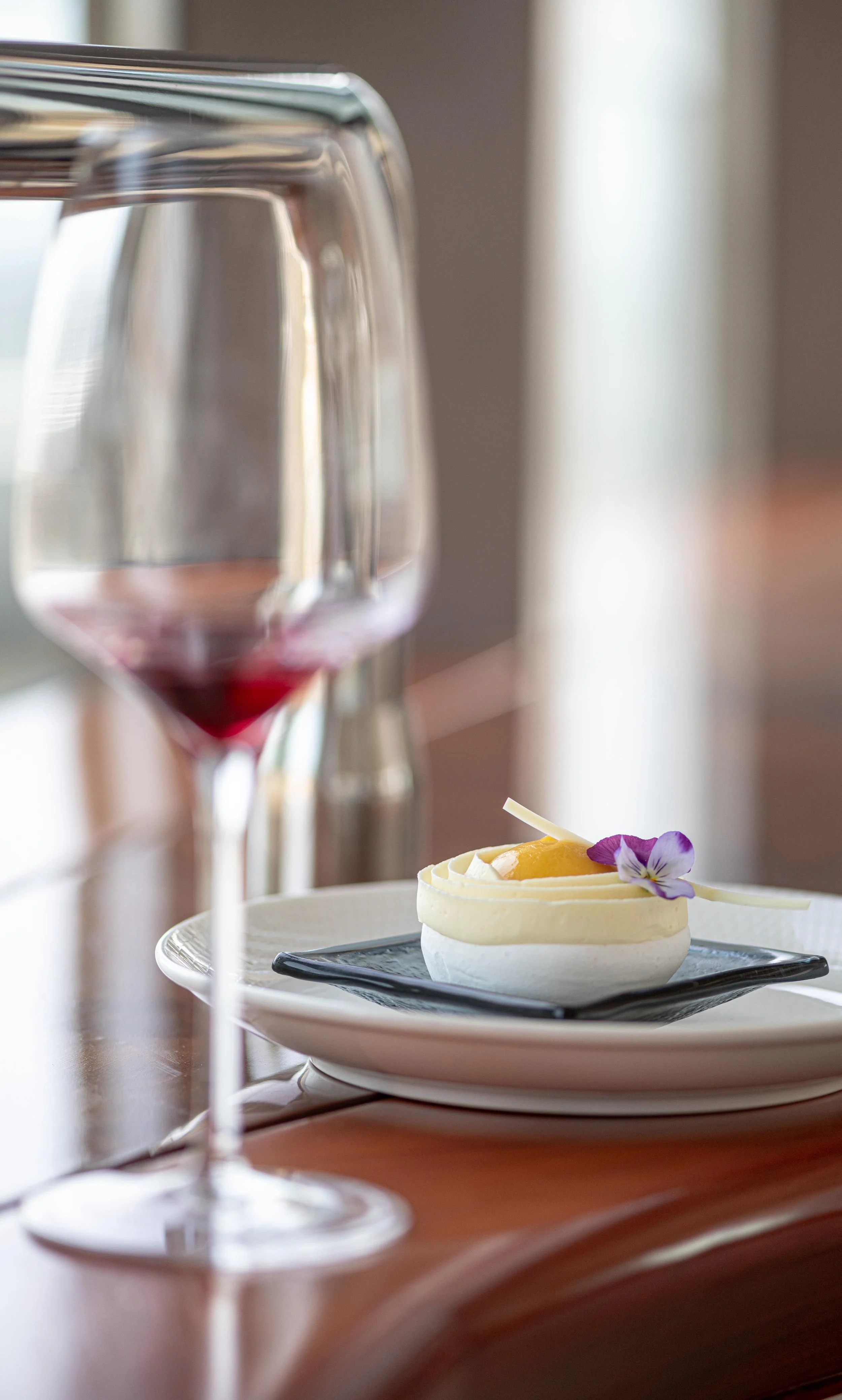 A dessert with cream, a sliced fruit, and an edible flower on top, served on a black square plate. A glass of red wine and a blurred background are visible.