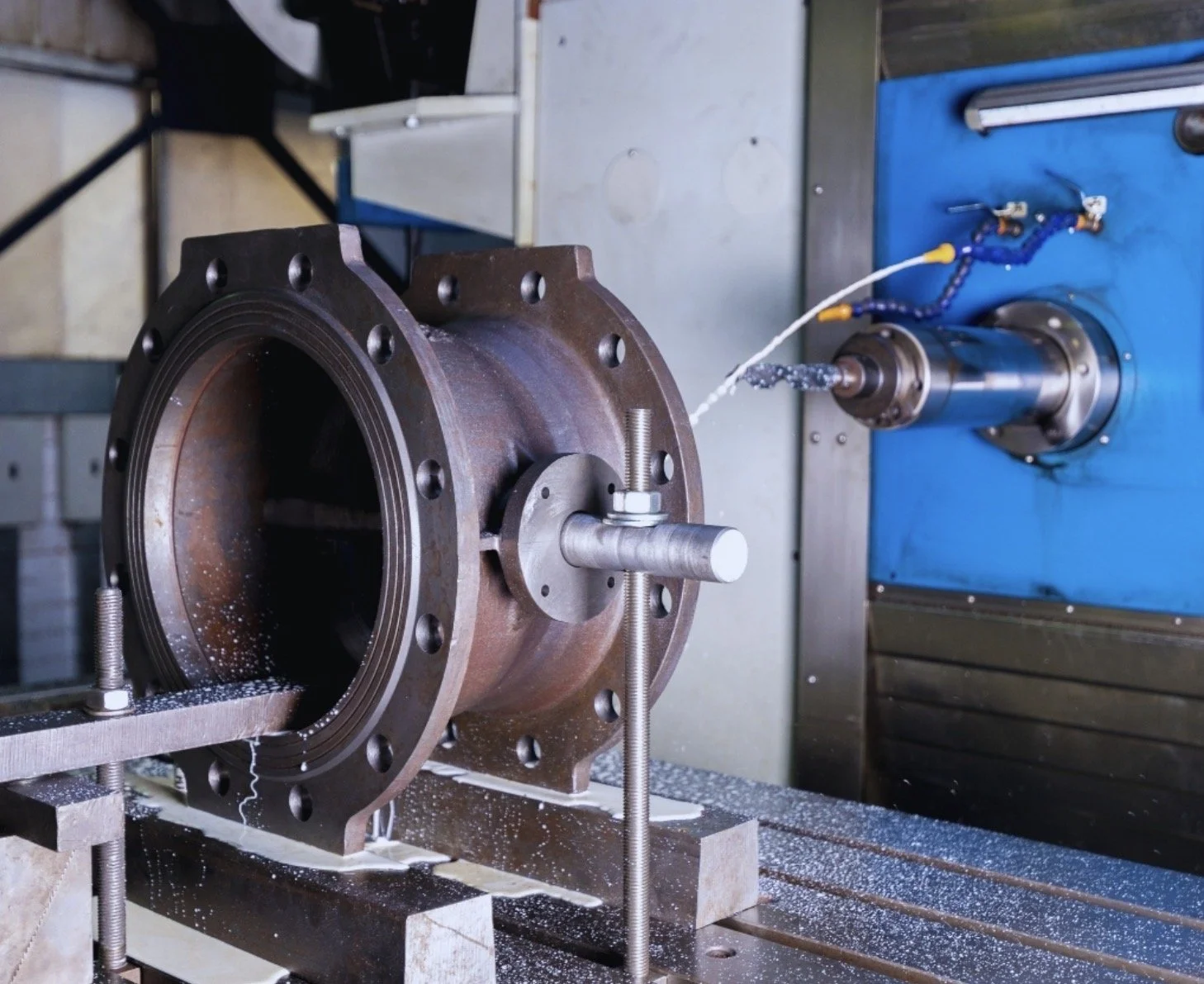 A large, rusty metal cylindrical component mounted on a machine bed, with a rotating cutting tool connected to a blue CNC machine in the background, engaged in a machining process with metal shavings and coolant fluid present.