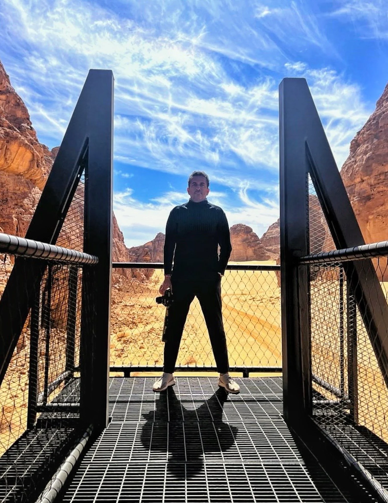 A man stands on a metal bridge with desert canyon walls in the background, under a blue sky with wispy clouds.