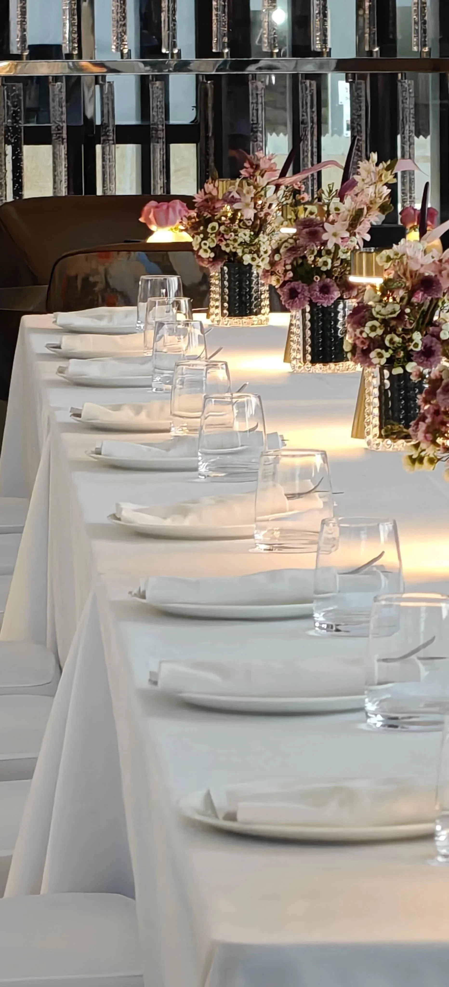 Elegant dining table set with white tablecloth, clear water glasses, white napkins, and floral centerpieces in vases with pink and white flowers.