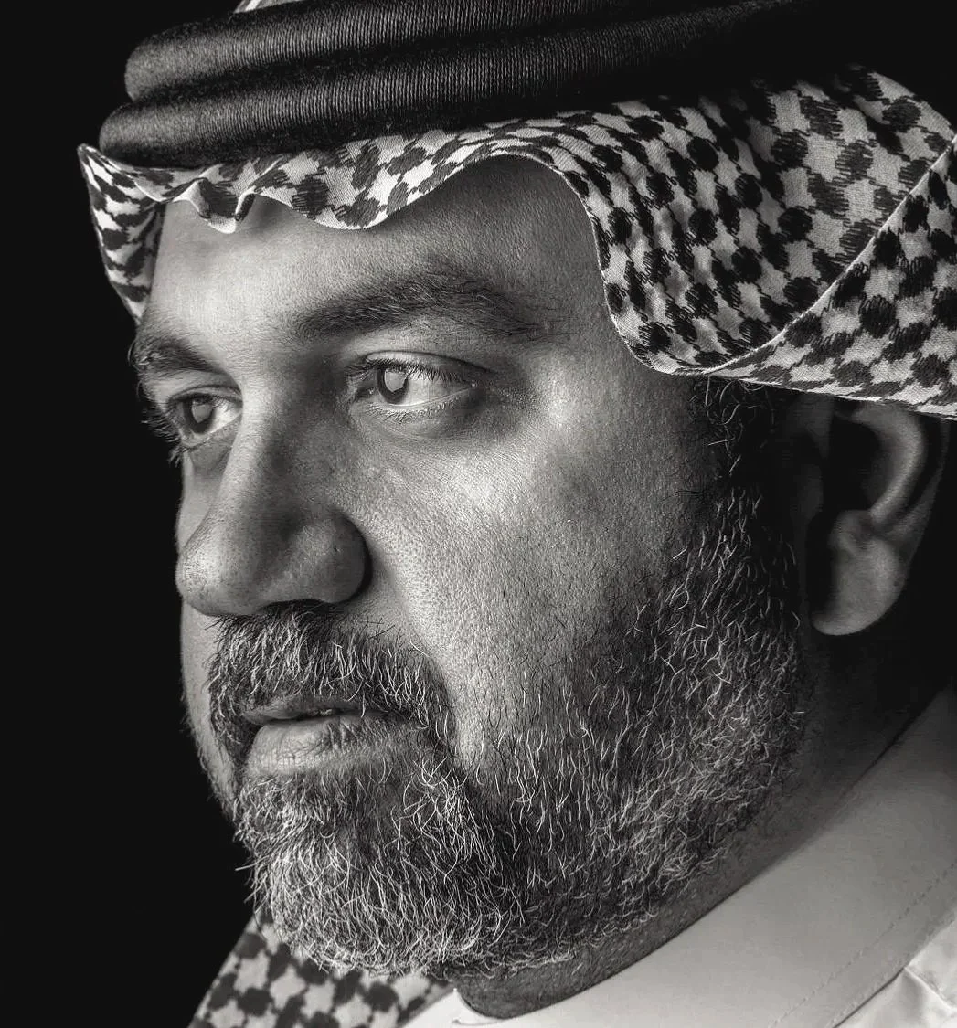 A black and white close-up portrait of a man wearing a traditional Middle Eastern headscarf, known as a keffiyeh or ghutra, with detailed patterns, and a thobe. The man has a full beard and a contemplative expression.