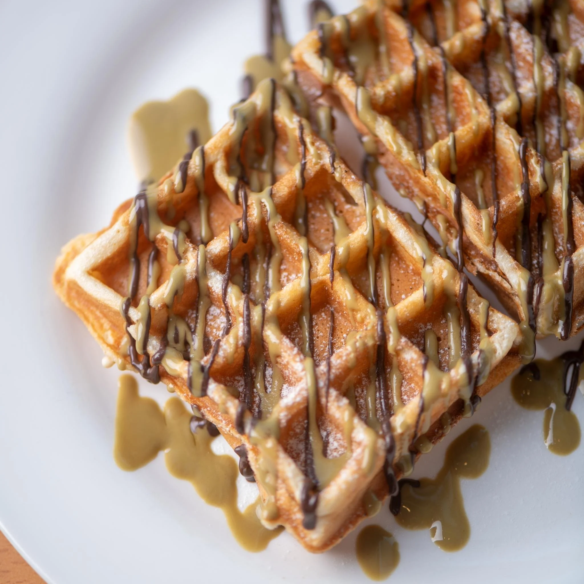 Close-up of a waffle with chocolate drizzle, caramel sauce, and light-colored icing on a white plate.
