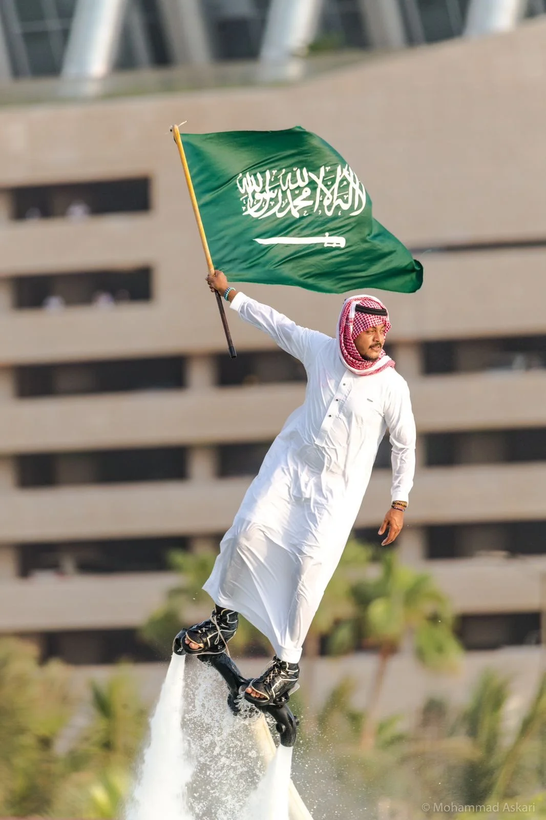 A man in traditional white Middle Eastern attire with a red and white headscarf rides a flyboard with a Saudi Arabian flag attached, against a background of a modern building.