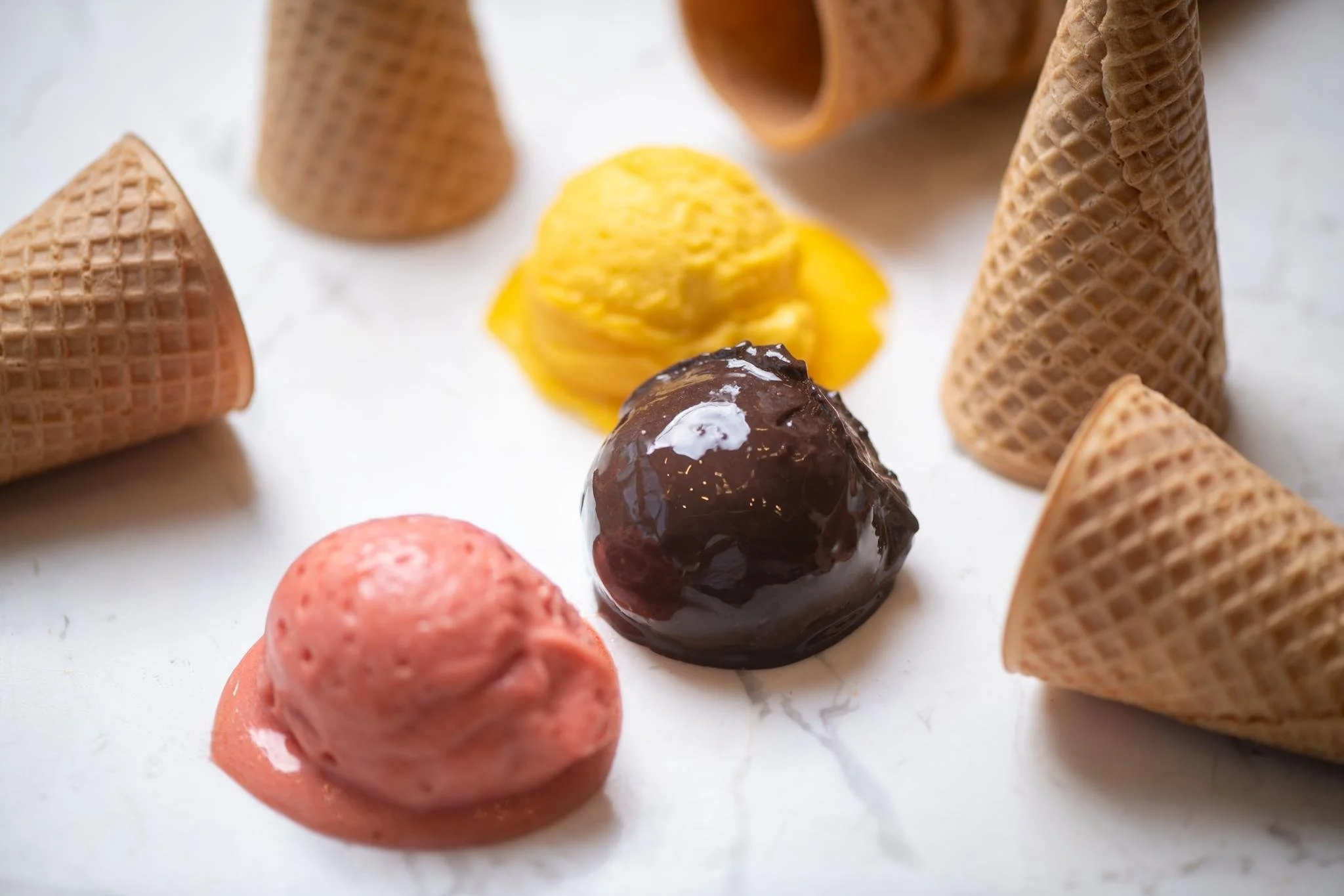 Close-up of various ice cream scoops including strawberry, chocolate, and vanilla, surrounded by waffle cones on a white surface.