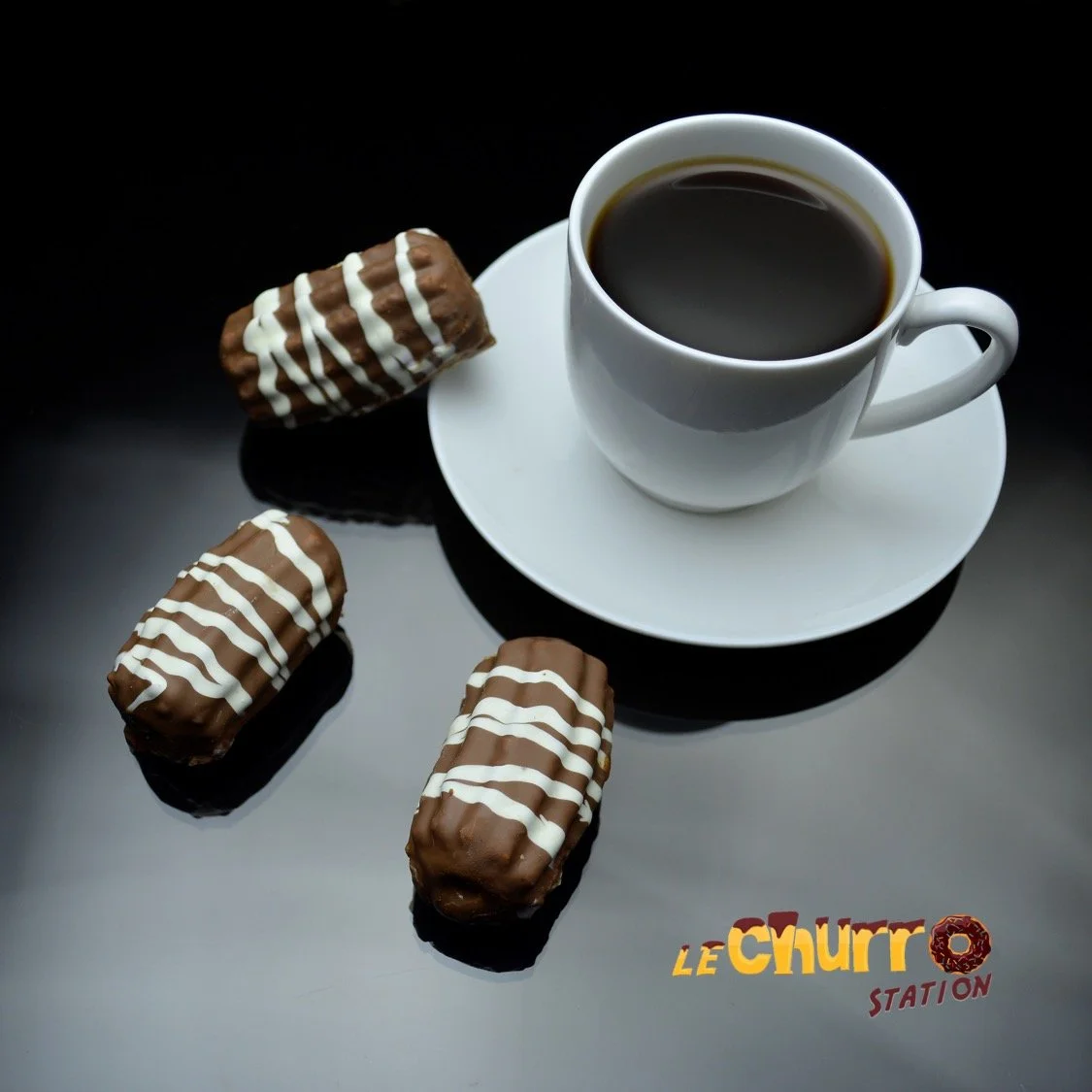 A white coffee cup filled with black coffee on a white saucer, placed on a black surface with three chocolate éclairs with white icing stripes nearby. The image has a logo at the bottom right that reads 'LE CHURR STATION'.