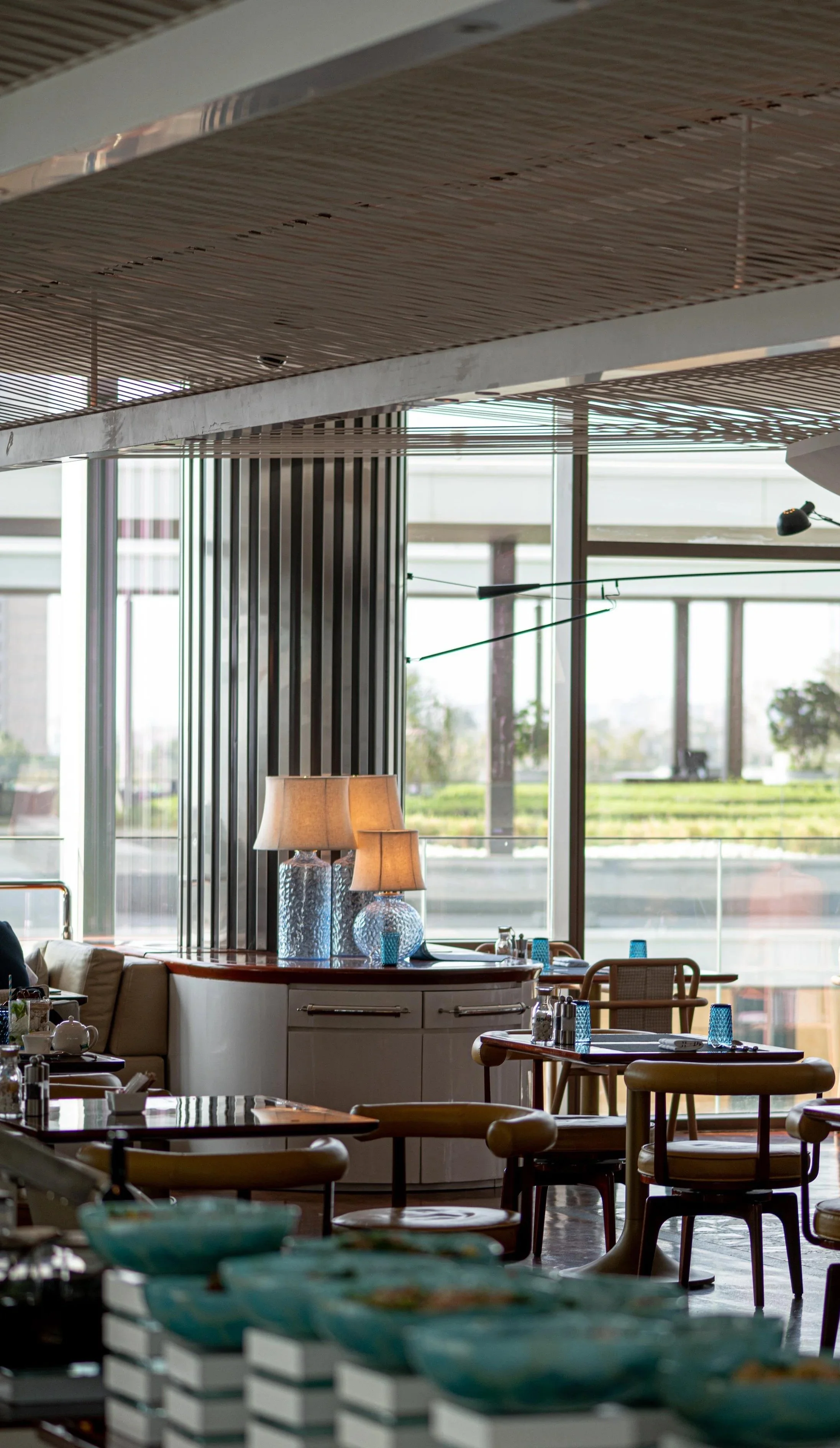 Interior of a modern restaurant or cafe with tables, chairs, and decorative lamps near large windows with a view of green outdoor space.