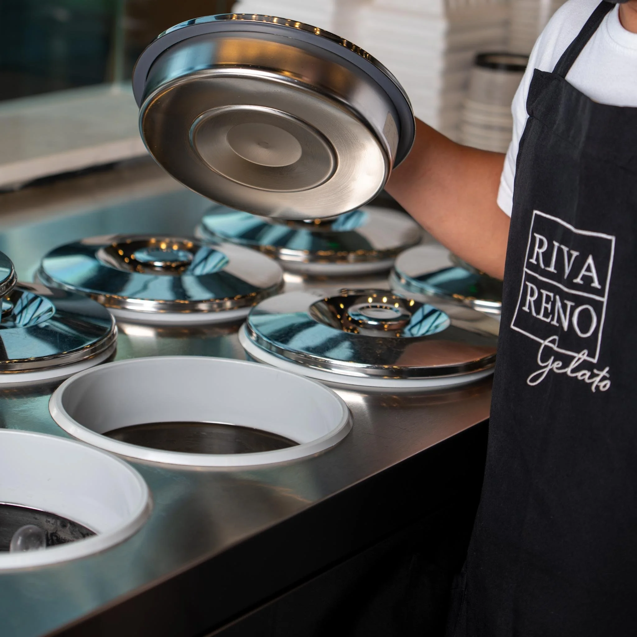Stainless steel and white open ice cream containers on a counter, with a person wearing a black apron and a white shirt nearby. The person is holding a metal container with a lid.