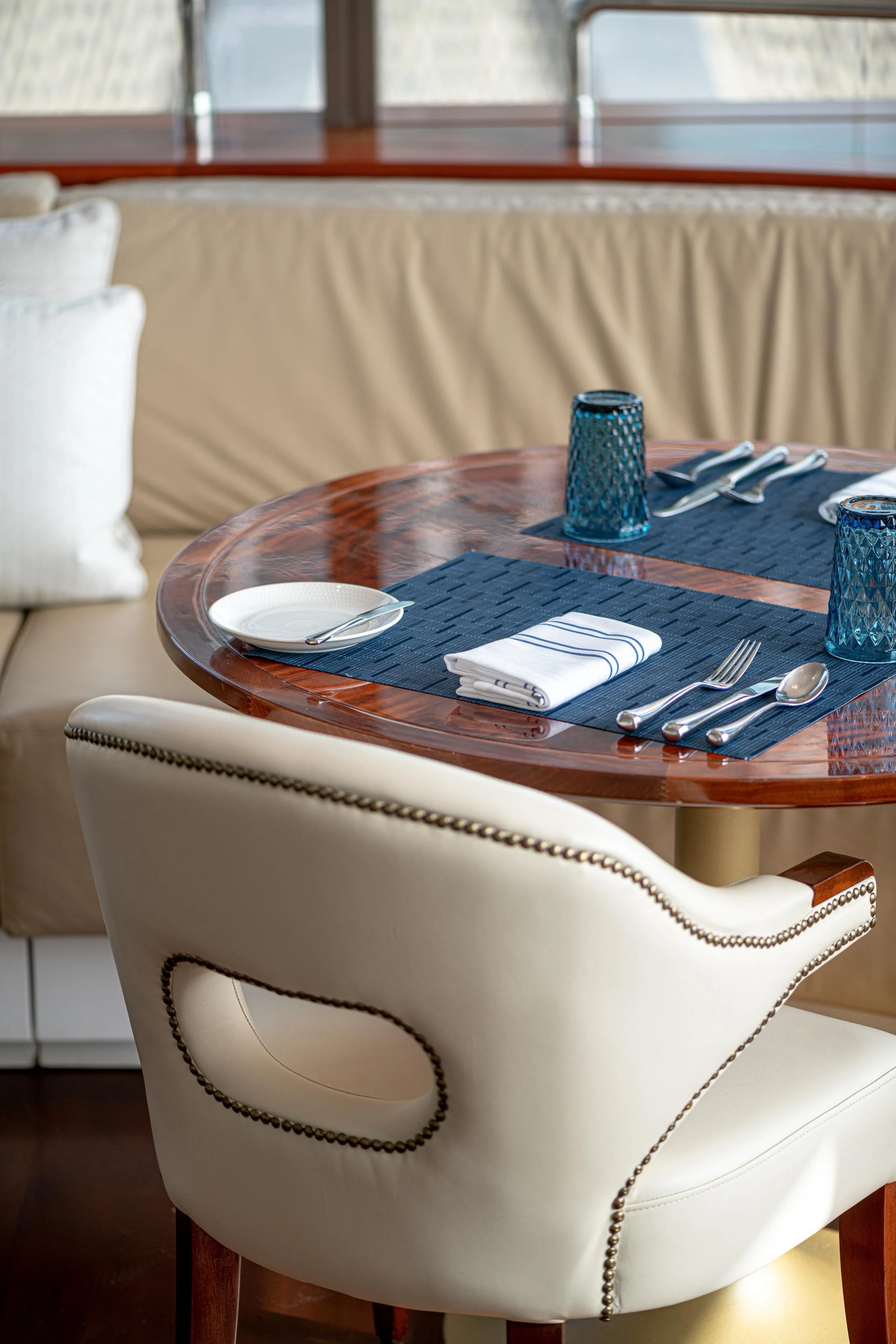 A round wooden dining table set with blue glasses, white plates, silver cutlery, and a white napkin with blue stripes, surrounded by white upholstered chairs with dark wooden legs and nailhead trim.