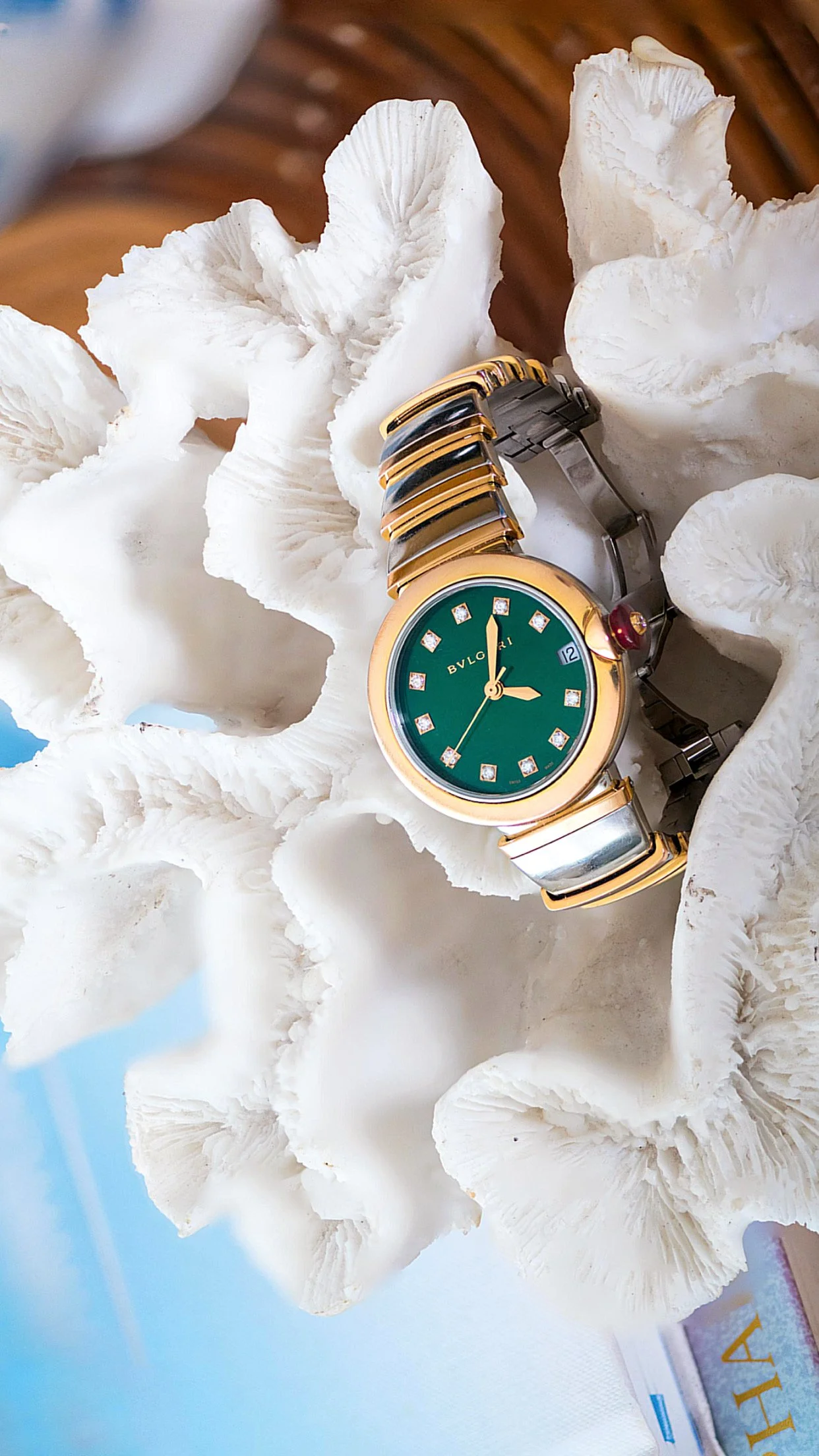 A luxury BVLGARI wristwatch with a green face, gold and silver band, and diamond hour markers, resting on white, textured coral-like rocks.