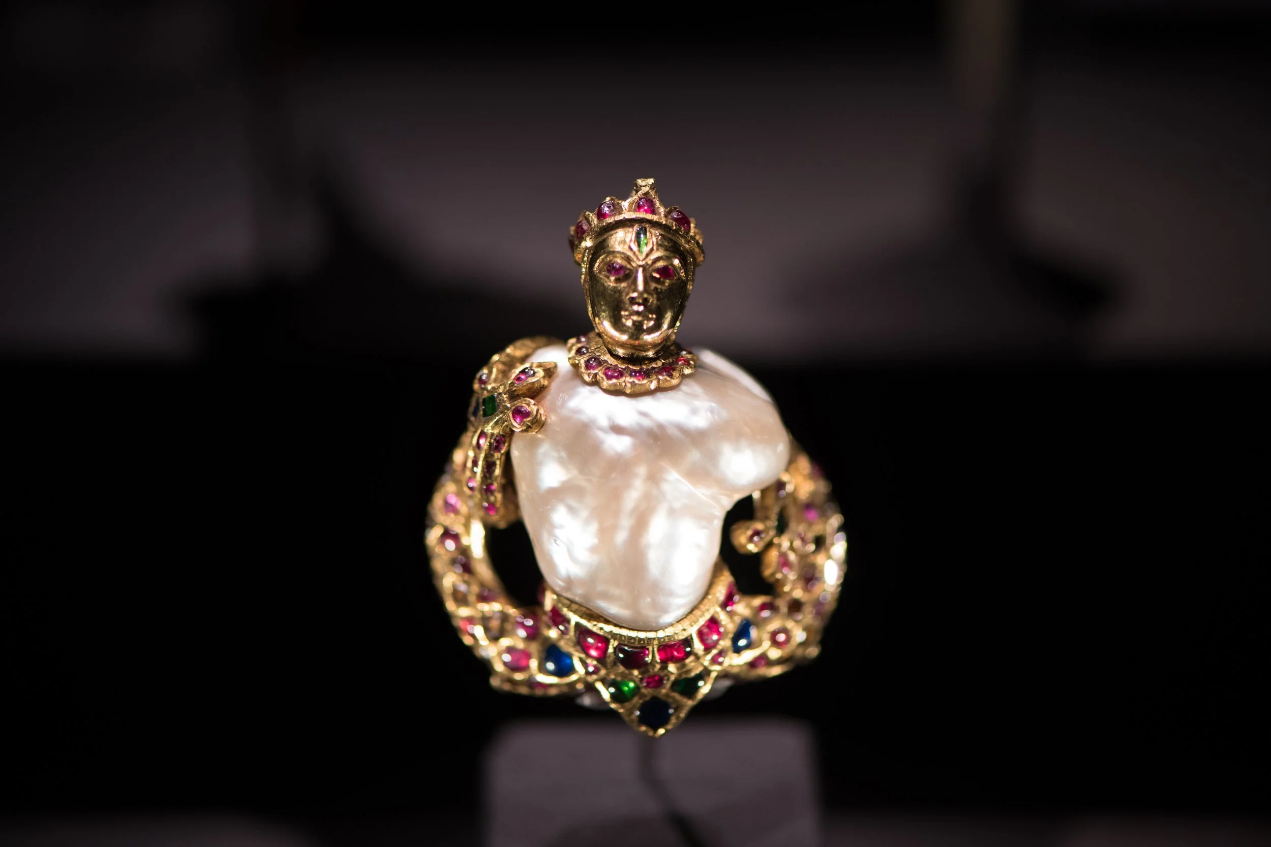 A jewelry piece featuring a figure of a female bust with a crown, adorned with colored gemstones and gold detailing, mounted on a pearl base.