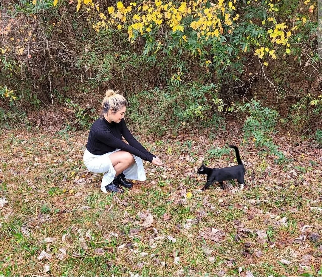 A woman crouching on the ground outdoors, wearing a black sweater and white striped skirt, playing with a black cat among fallen leaves and greenery.