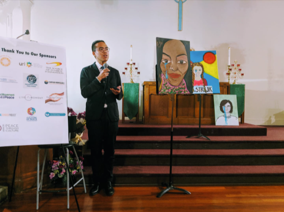 A man in a suit speaking at a church event, with colorful artwork and a banner of sponsor logos behind him.
