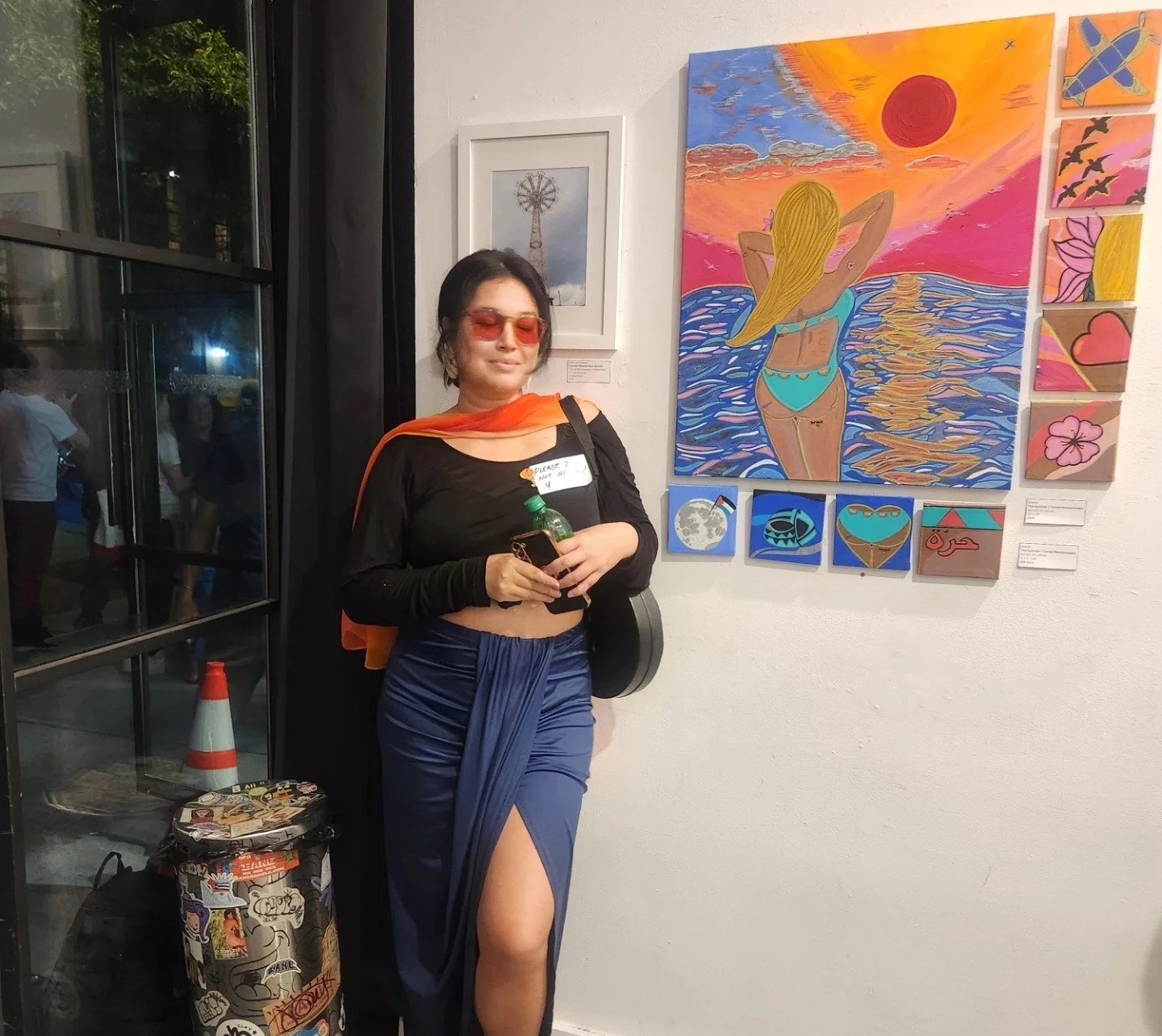 A woman with short dark hair wearing a black top with a sheer overlay, orange sunglasses, blue skirt with a thigh-high slit, and a sheer orange scarf draped over her shoulders, standing in an art gallery next to colorful paintings including a large a