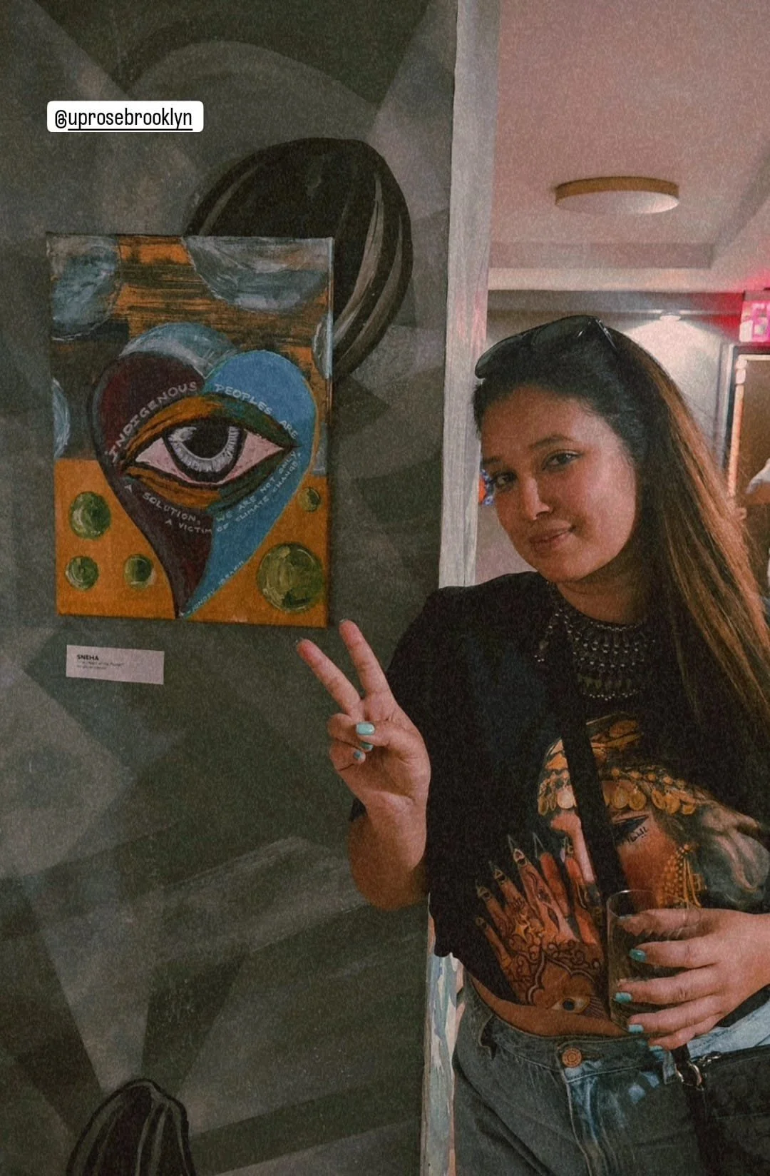 A woman with brown hair wearing sunglasses on her head, a black shirt, and a colorful tattoo on her arm, making a peace sign and holding a drink, stands next to an abstract painting featuring an eye and a heart.