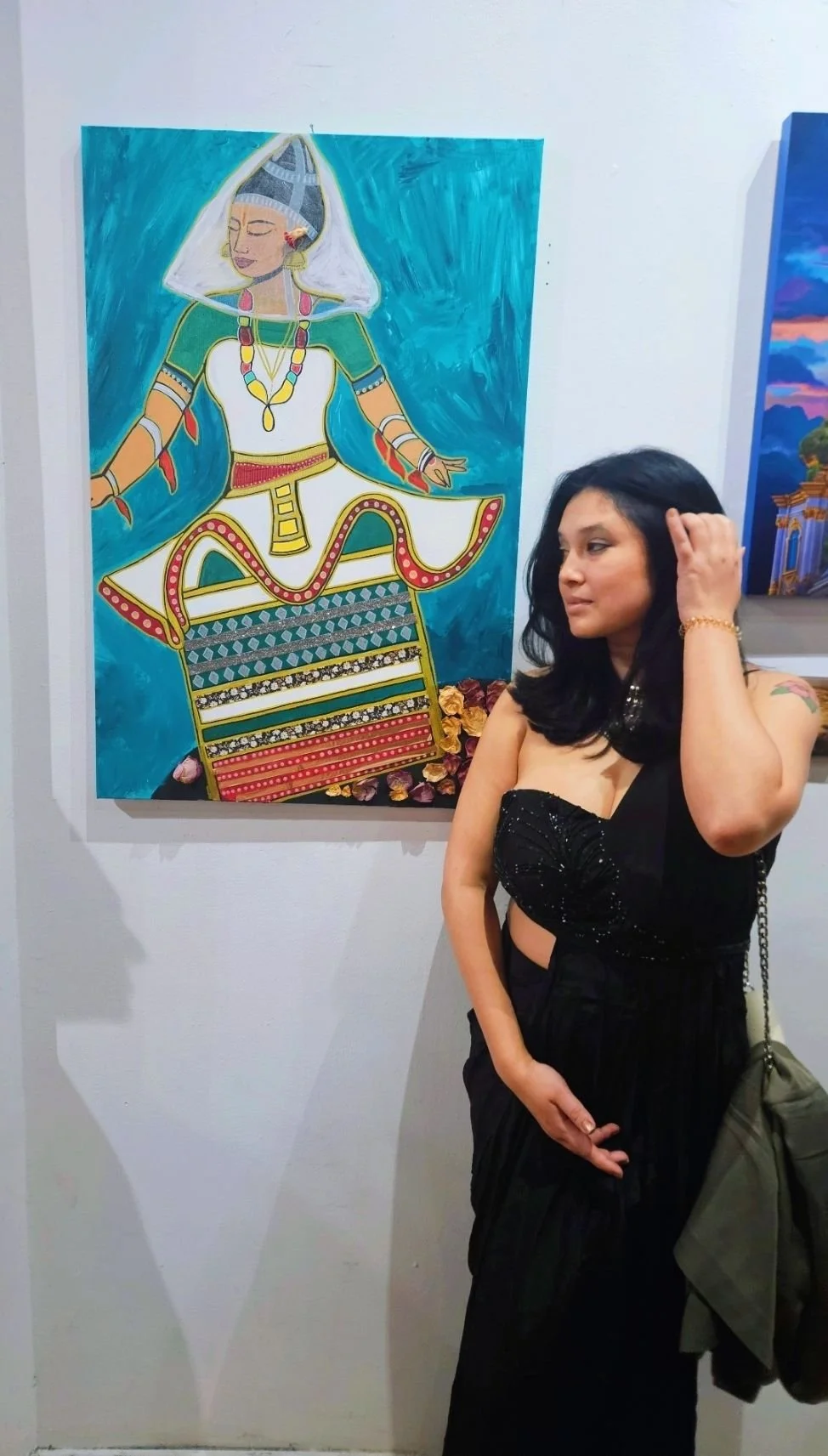 A woman with black hair, wearing a black top and pants, stands in front of a colorful painting of a woman in traditional attire, sitting on a decorated platform, with blue background. The woman is touching her hair and looking at the painting.