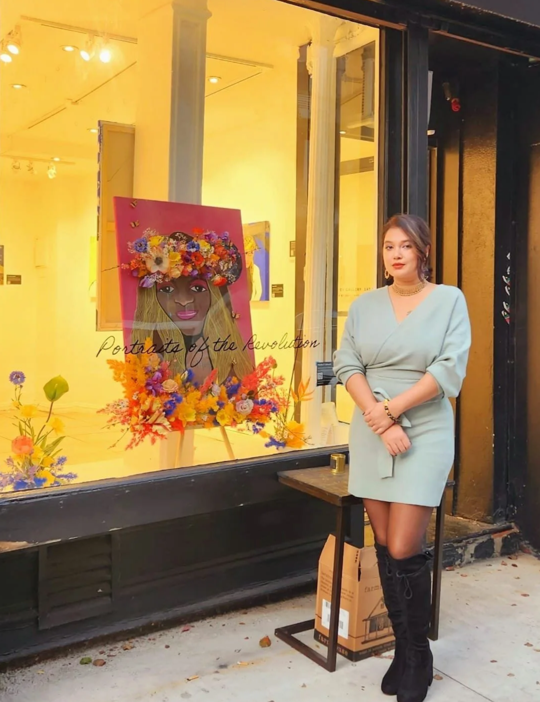 A young woman standing outside a gallery window featuring a colorful portrait of a woman with a flower crown, surrounded by vibrant flowers, with the text 'Portraits of the Revolution' on the window.