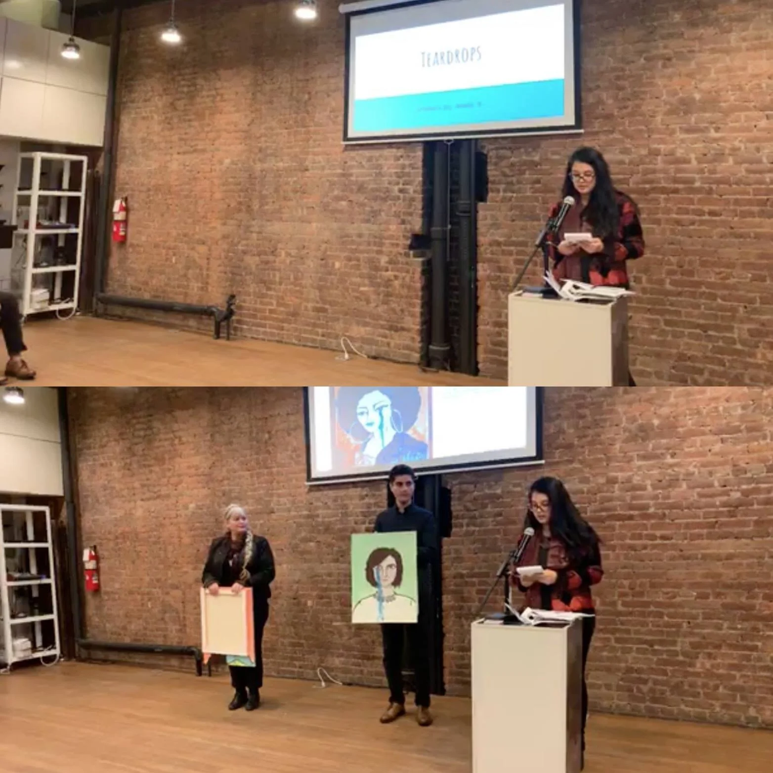 A group of people participating in a presentation or event in a room with a brick wall background and a large screen displaying artwork. The attendees are holding artwork or posters, and a woman at a podium is reading from a paper.