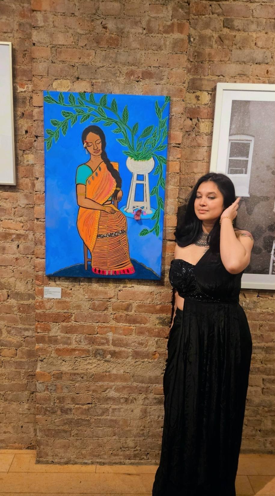 A woman with long black hair wearing a black, strapless dress stands next to a colorful painting of a woman in traditional Indian attire, sitting with her eyes closed, surrounded by green foliage and a white pot on a stand, on a brick wall background
