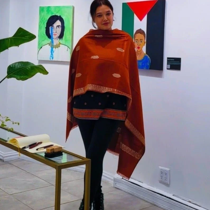 A woman standing in an art gallery with two paintings on the wall behind her, one featuring a woman with black hair and a tear, and the other with abstract geometric shapes. She is wearing a red and orange shawl, black pants, and black shoes.