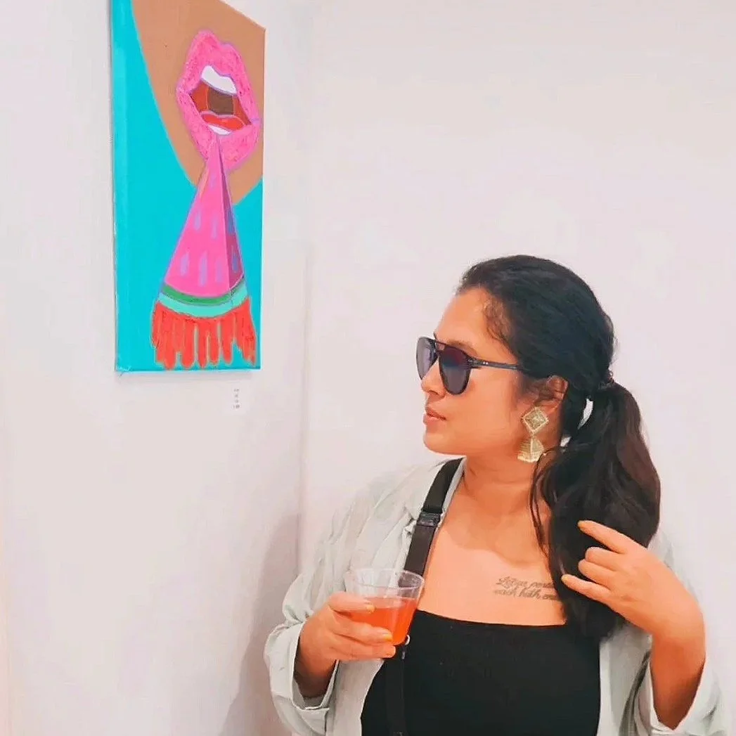 A woman with dark hair, sunglasses, and earrings looks at a colorful abstract art piece of lips and tongue on a wall in an art gallery, holding a drink in her hand.