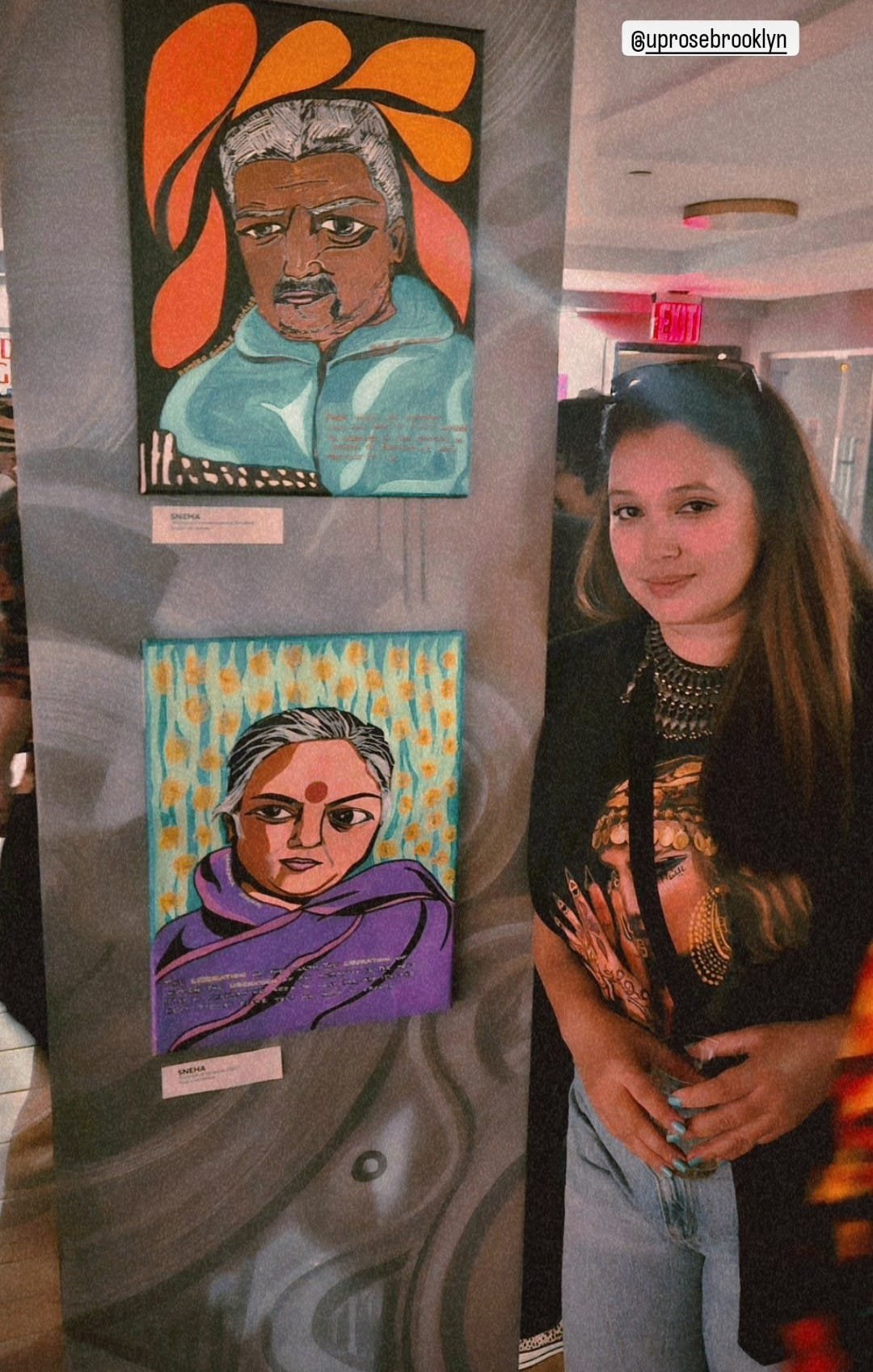 A woman standing next to a display of two colorful portraits of women. The portraits are painted in a stylized, expressive manner, with vibrant backgrounds. The woman has long hair and is wearing a black shirt with jewelry. An Instagram handle '@upro