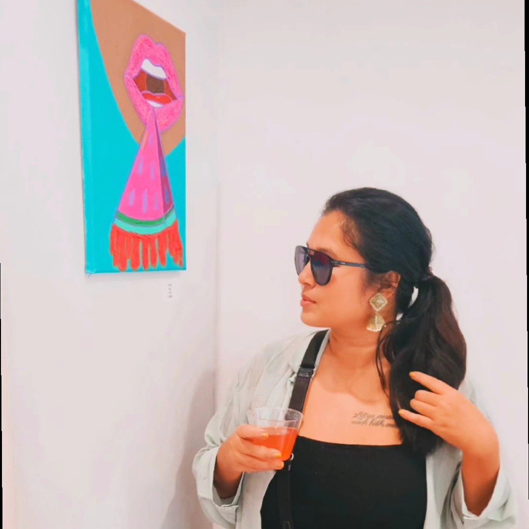 A woman with dark hair, sunglasses, and earrings is looking at an artwork on a white gallery wall. She is holding a drink and has a tattoo on her chest. The artwork features a pink and red stylized mouth with an extended tongue.