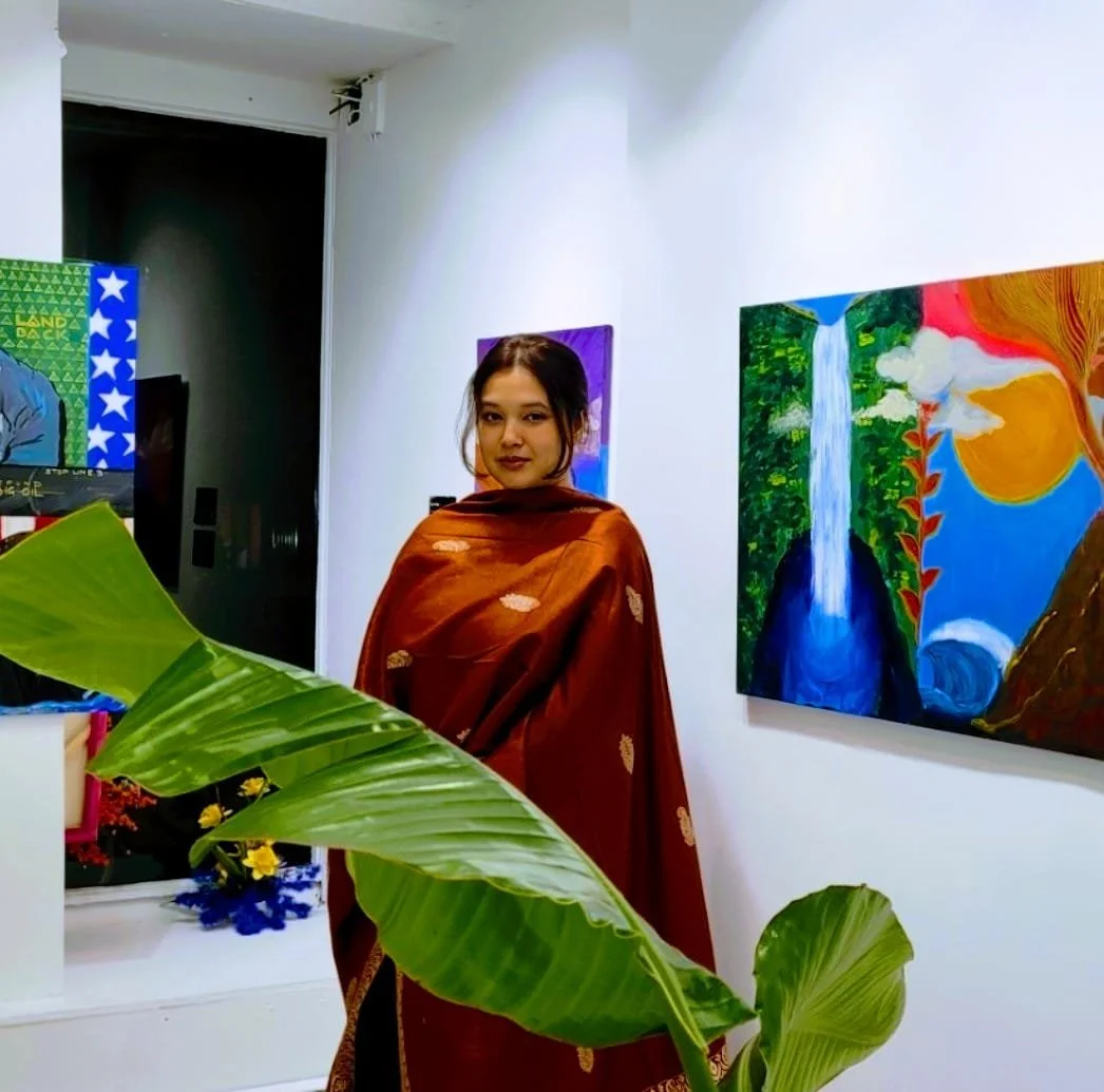 A young woman wearing a brown sari standing in an art gallery with colorful paintings on the white walls and large green leaves in the foreground.