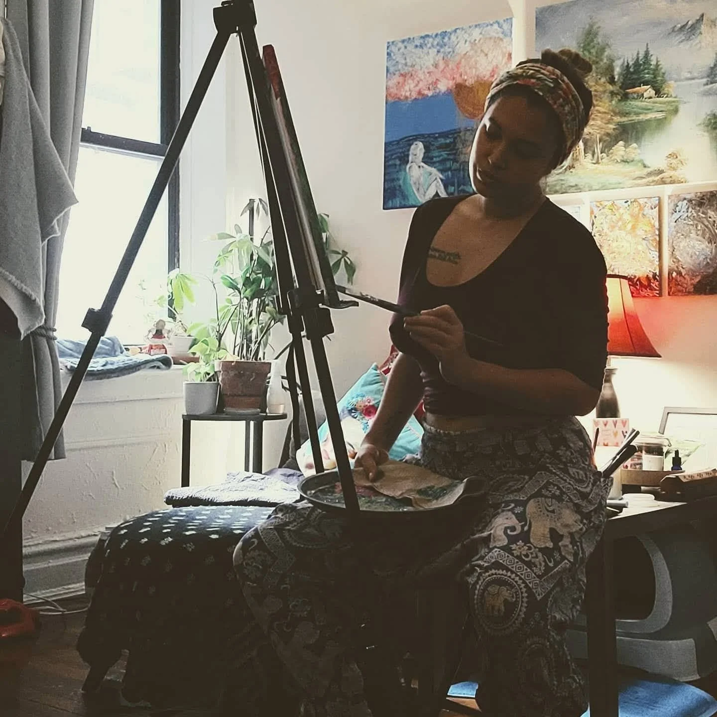 A woman with a colorful headscarf and patterned pants is sitting in front of an easel, working on a painting in a well-lit room decorated with landscape paintings and houseplants.