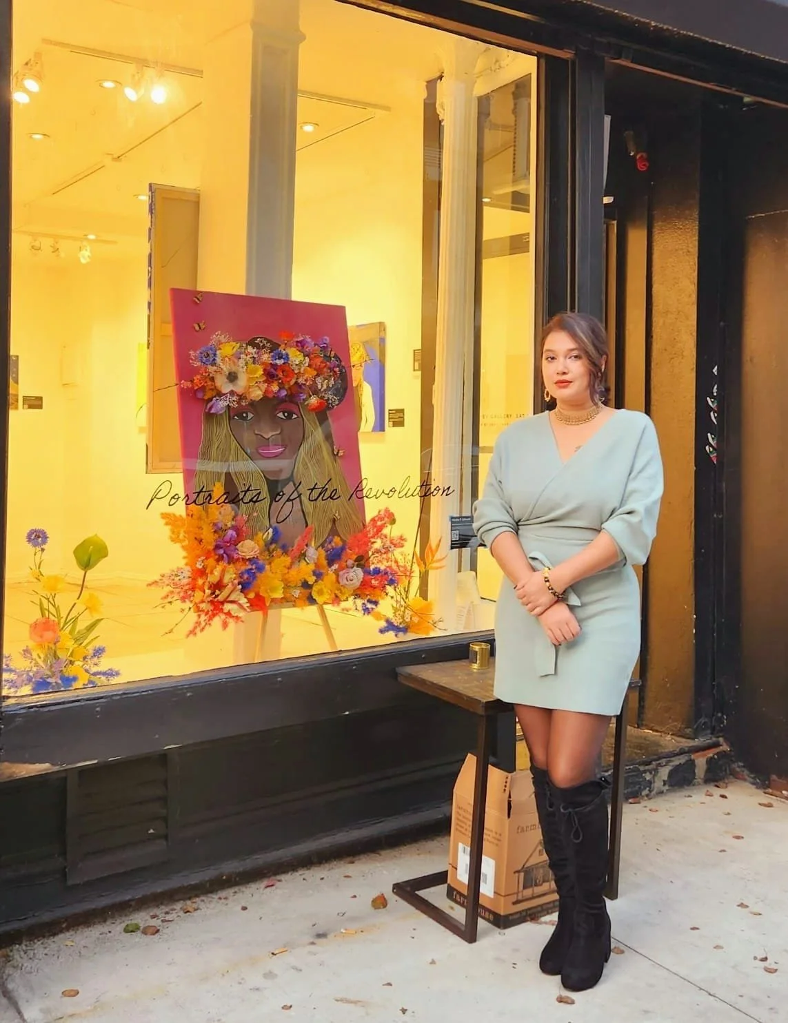 A woman in a light green dress and black knee-high boots stands outside an art gallery with a glass window. Inside the window, there's a colorful portrait of a woman with a floral crown, surrounded by vibrant flowers, with the words 'Portraits of the