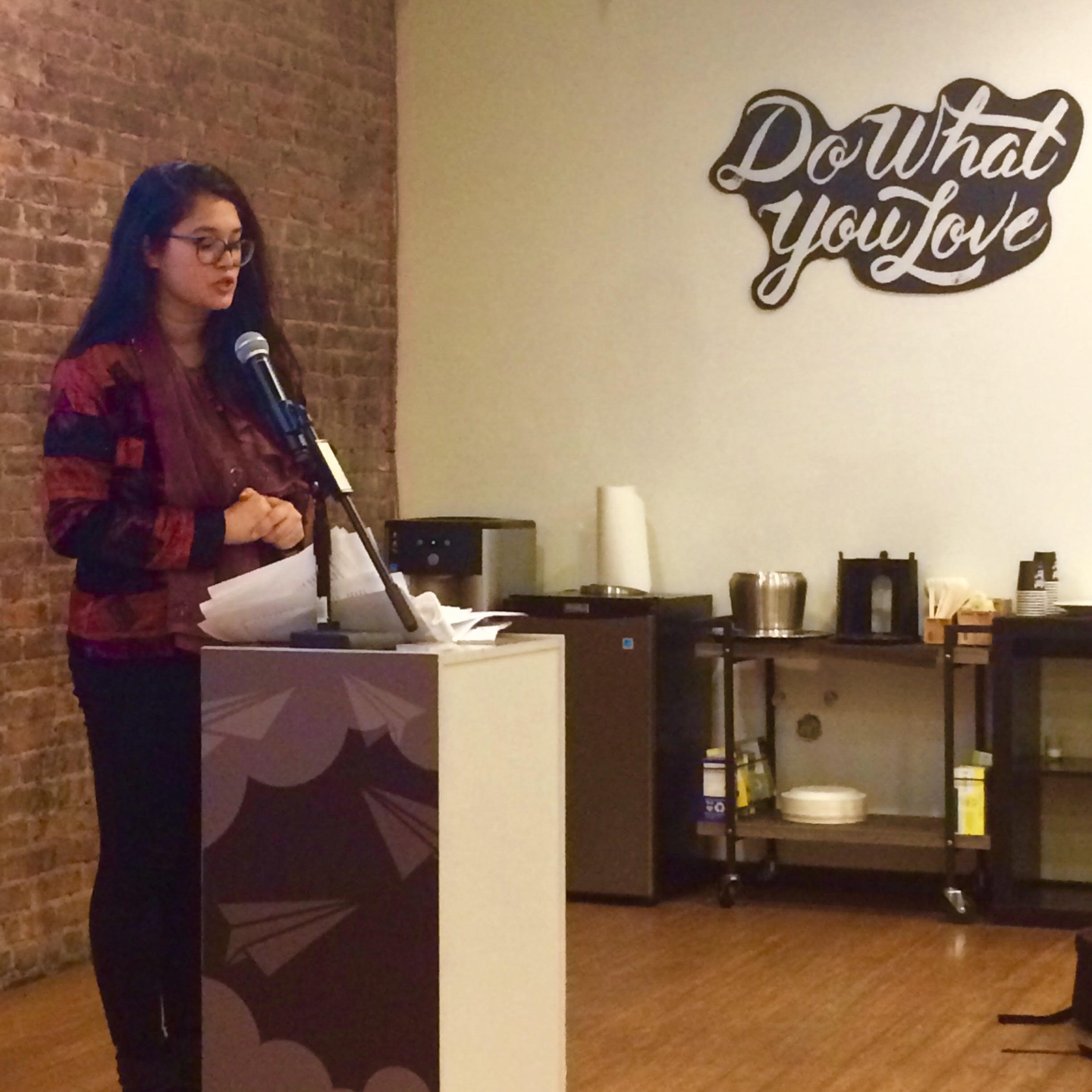 A woman with long dark hair and glasses is standing behind a podium with papers in her hands, speaking into a microphone. The background features a brick wall on the left and a cream-colored wall on the right with a decorative sign that reads 'Do wha
