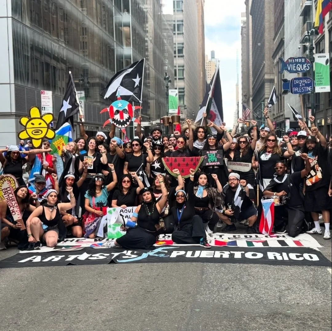 A diverse group of protesters gathered on a city street, holding flags and signs, with some raising their fists. The crowd is behind a large banner that reads 'Artistas y Puertorico.' The participants are expressing solidarity and activism, with some