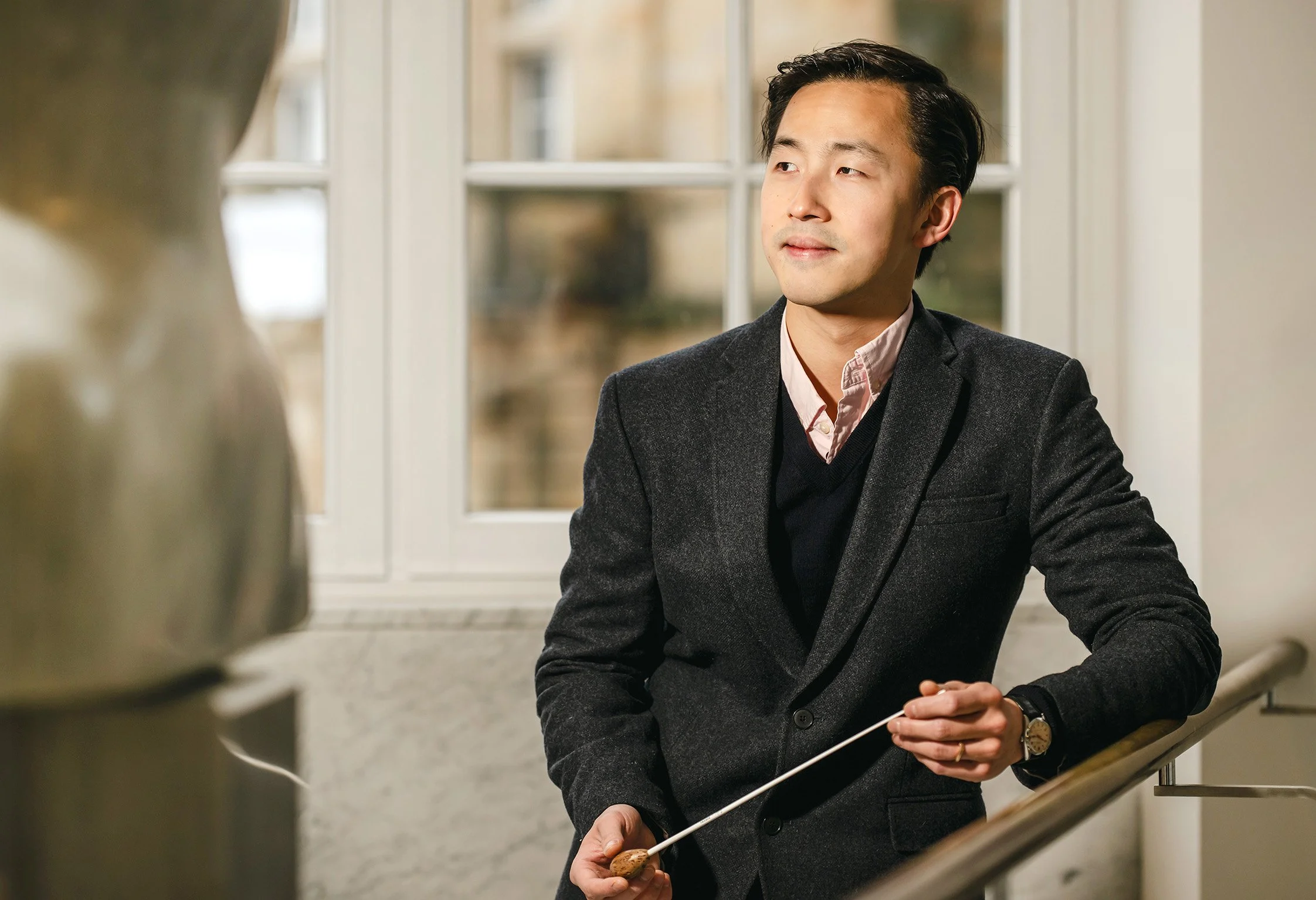 Man in blazer and dress shirt sitting on staircase, looking to his right, holding an object in his right hand with a white stick or pointer, near a large window in a well-lit indoor space.